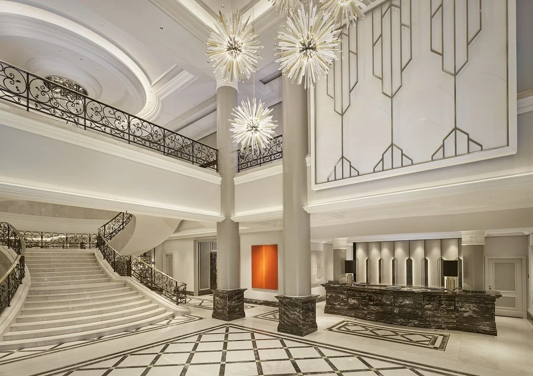 Elegant hotel lobby with marble floors, large columns, floral chandeliers, a curved staircase, and a reception desk.