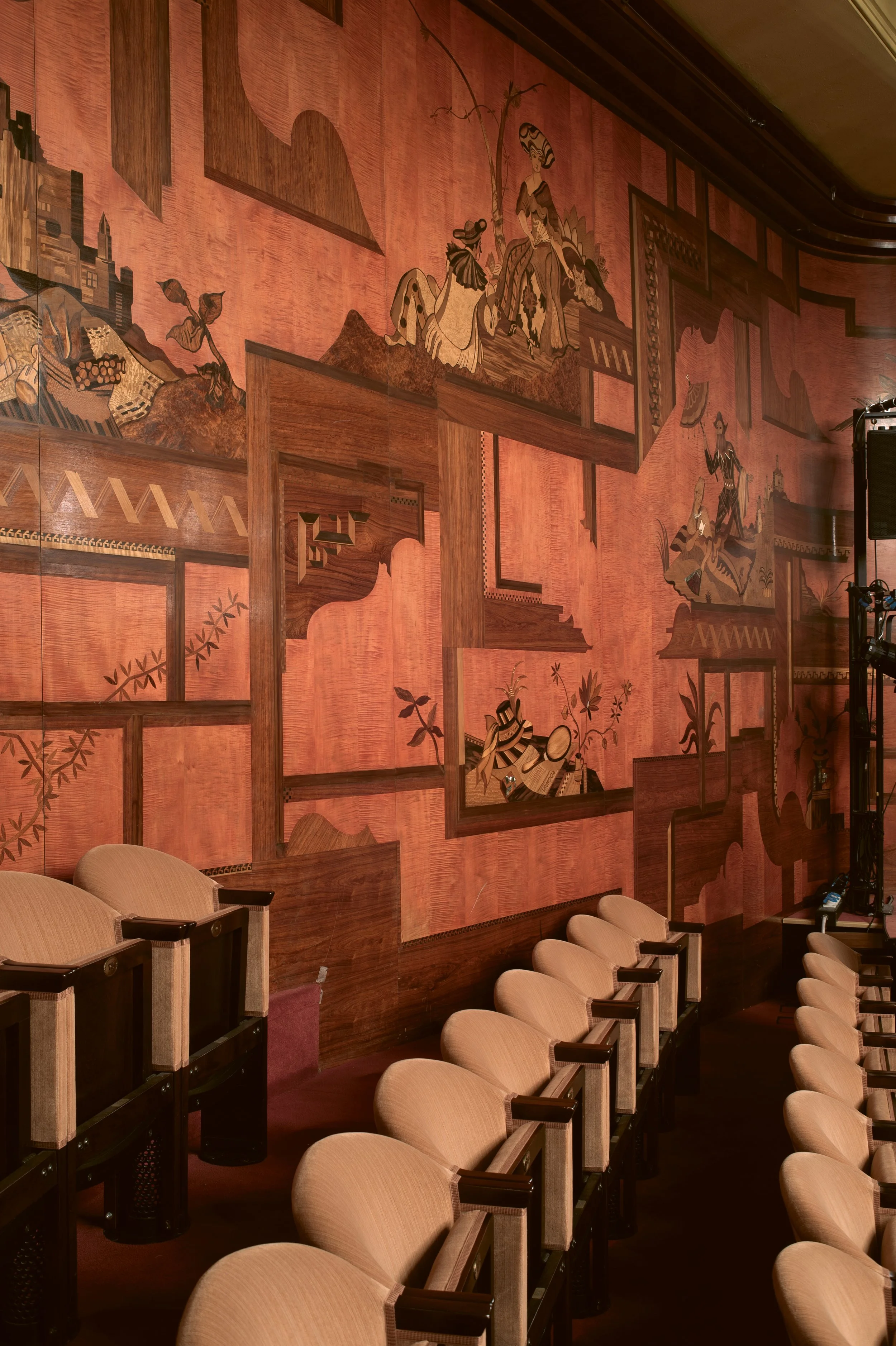 The photograph shows a row of beige theater or auditorium seats with dark wooden armrests, situated in front of a large decorative wooden wall mural. The mural features intricate artwork with figures in traditional Asian attire, positioned among geometric and nature-inspired patterns, creating a rich, detailed background.