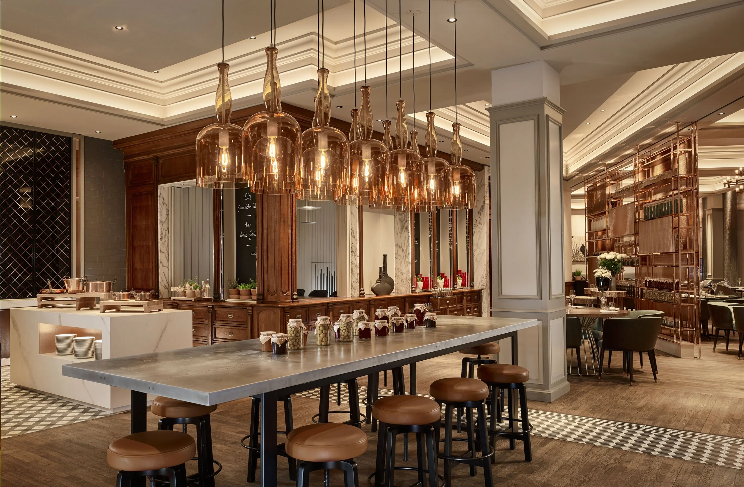 Elegant restaurant interior with a modern bar area featuring hanging amber glass pendant lights, a marble-topped counter, and a spacious dining area with black chairs, decorative plant arrangements, and a partition with copper accents.