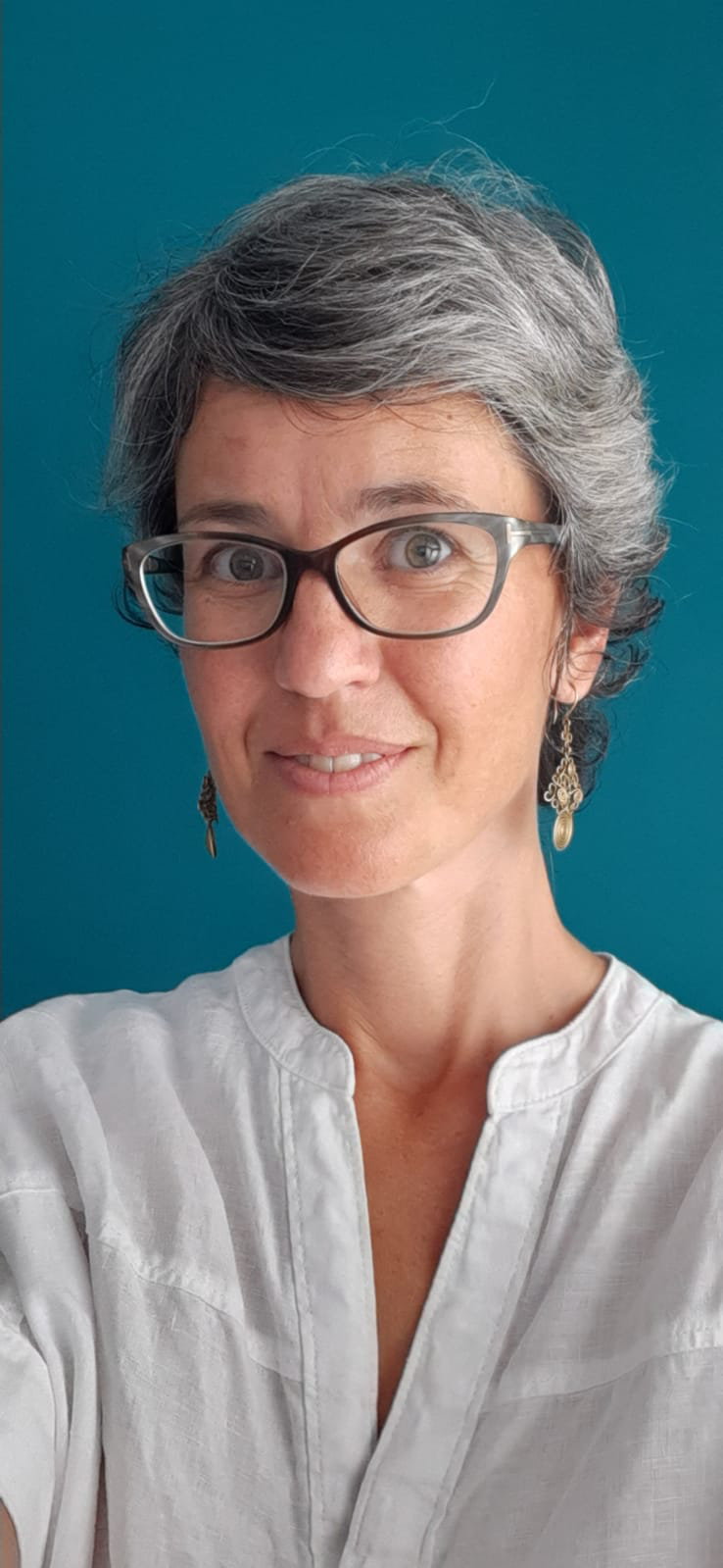 A woman with short gray hair, wearing glasses and earrings, wearing a white blouse, smiling in front of a teal background.
