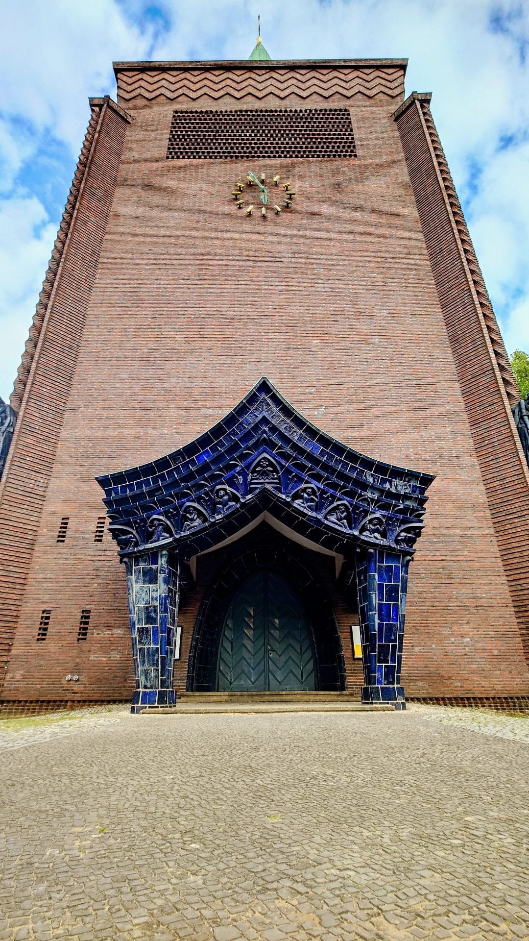 A tall brick church building with a clock, decorative blue mosaic archway at the entrance, and a clock showing 12:00, under a partly cloudy sky.