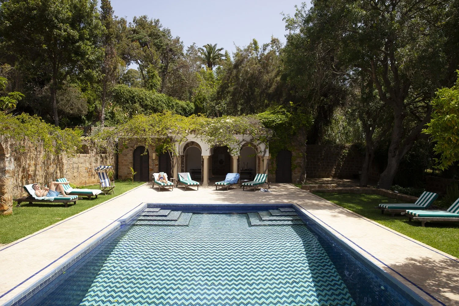 A backyard with a swimming pool, surrounded by lounge chairs, trees, and a stone wall. A person is lying on one of the lounge chairs.