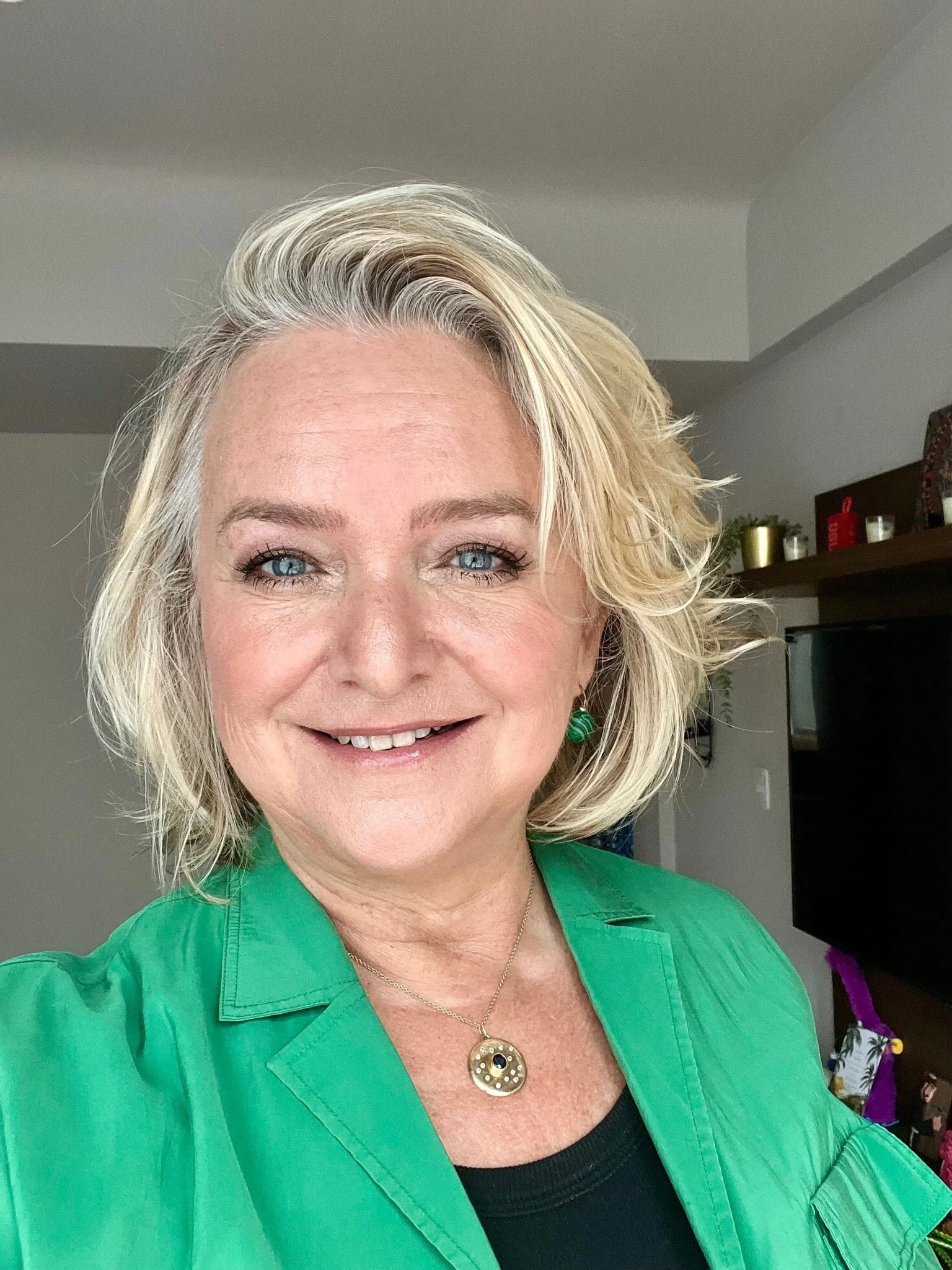 A woman with blonde hair and blue eyes smiling, wearing a green blouse and gold jewelry, in a warmly lit indoor setting.