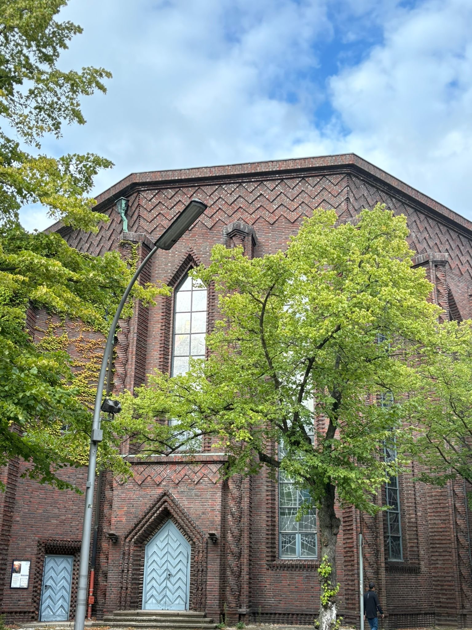 Red brick church with arched windows and a wooden door, surrounded by green trees and a street light, under a partly cloudy sky.