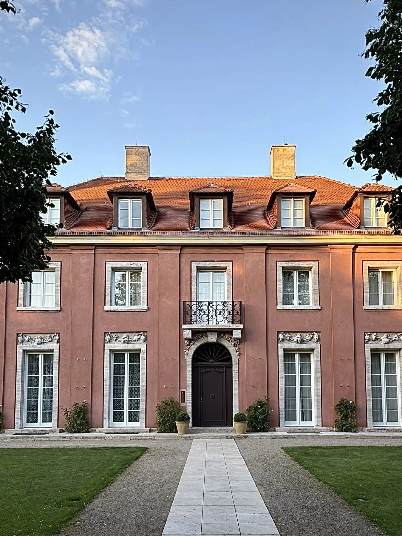 A large pink mansion with white trim, multiple windows, and a black front door, surrounded by a well-manicured lawn and tall trees.