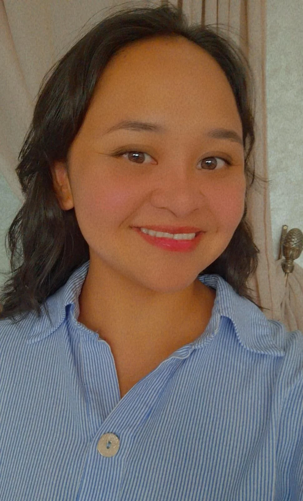 A woman with shoulder-length dark hair, wearing a light blue striped shirt, smiling in front of a patterned wooden background.