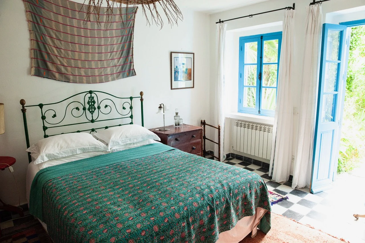 A cozy bedroom with a metal bed frame, green bedspread, and white pillows. There is a wooden nightstand with a lamp and decor, next to a window with bright blue shutters and white curtains. An open door to the outside reveals greenery.