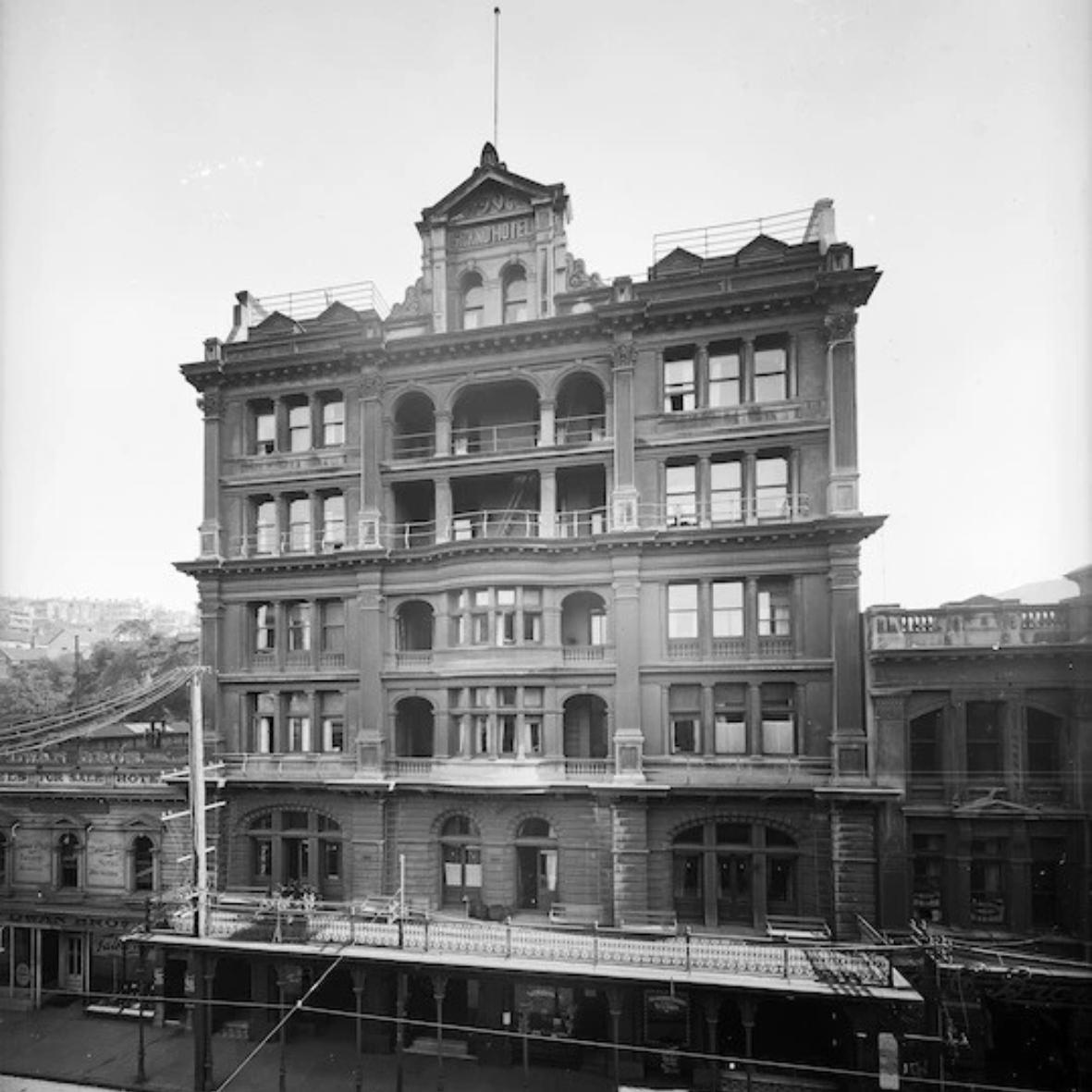 A historic black and white photograph of a multi-story building labeled as a hotel, featuring ornate architectural details, balconies, and a rooftop flagpole, with a street in the foreground.