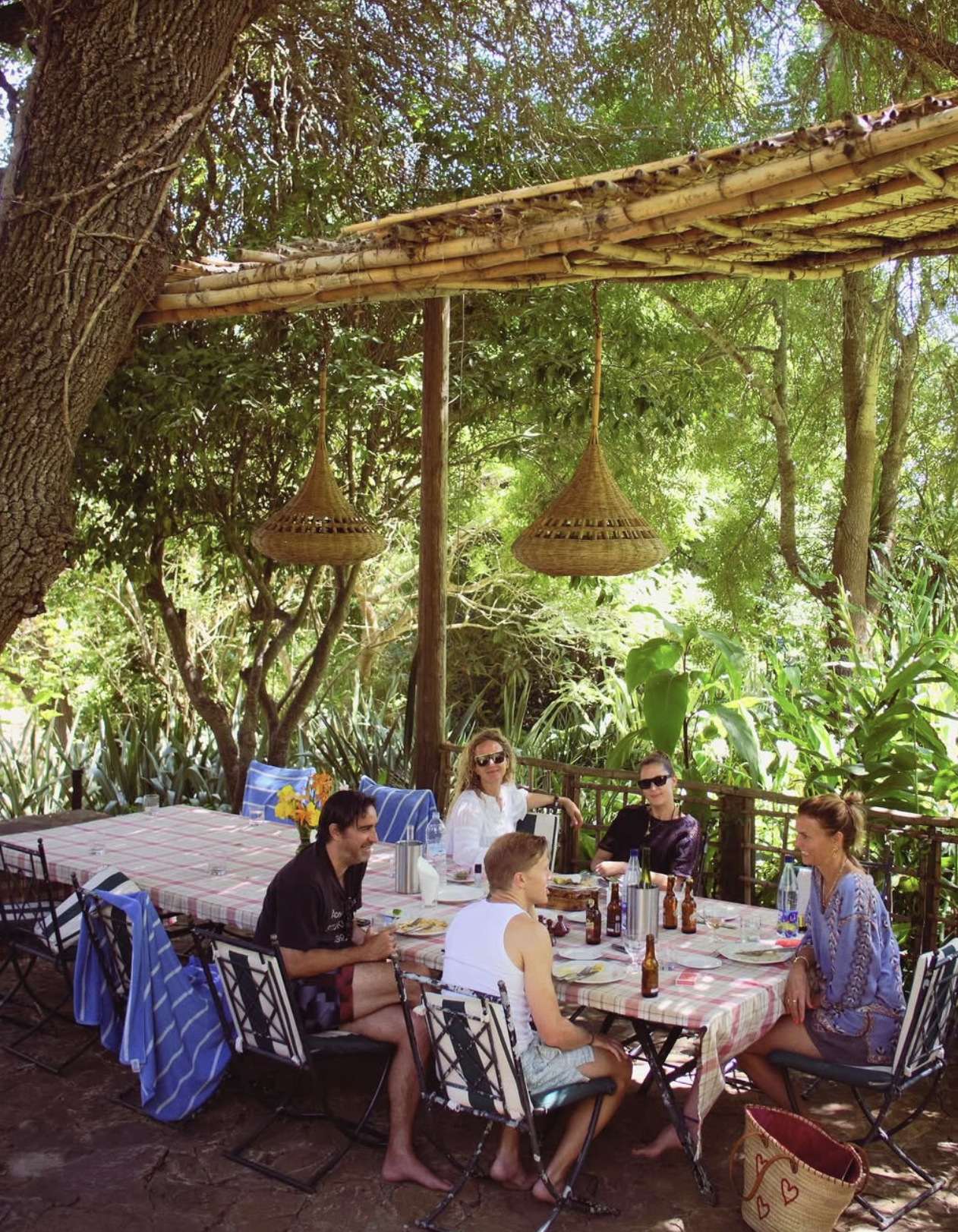 A group of six people sitting around a rectangular table outdoors in a lush, green garden. The table has a red and white checkered tablecloth, and there are bottles, glasses, and plates on it. Two women and four men are engaged in conversation, with some wearing sunglasses. The setting features dense foliage, trees, and hanging wicker lamps overhead.
