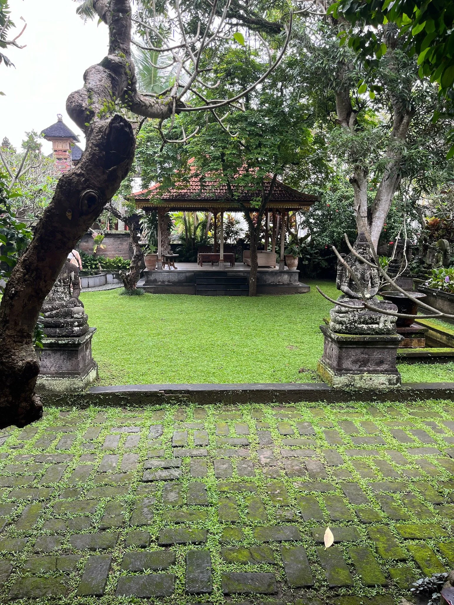 A lush garden with green grass, trees, and statues, featuring a wooden pavilion in the background and an moss-covered cobblestone path in the foreground.
