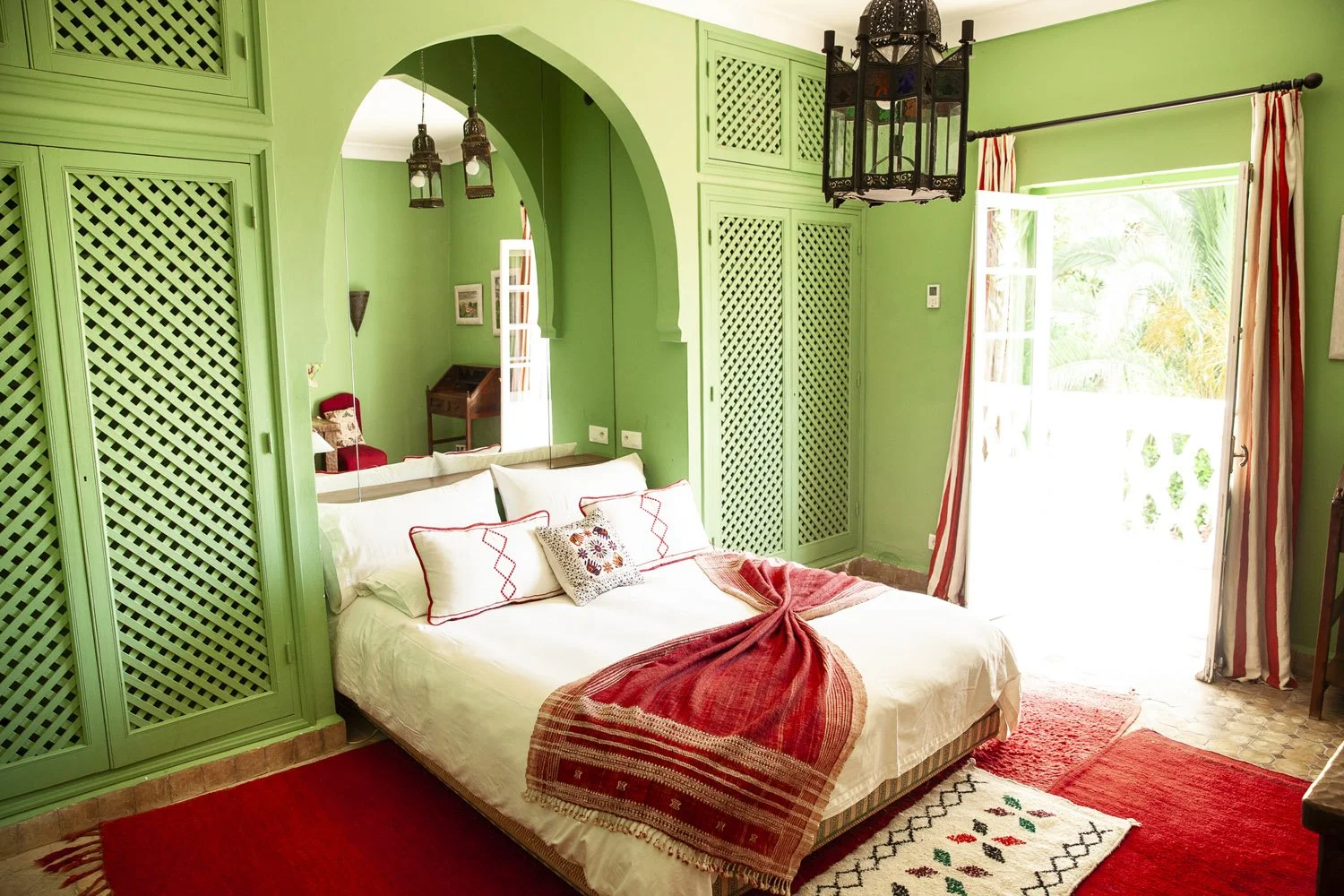Bright bedroom with green walls, a bed with white linens and red accents, and an open balcony door with striped curtains, surrounded by tropical outdoor greenery.