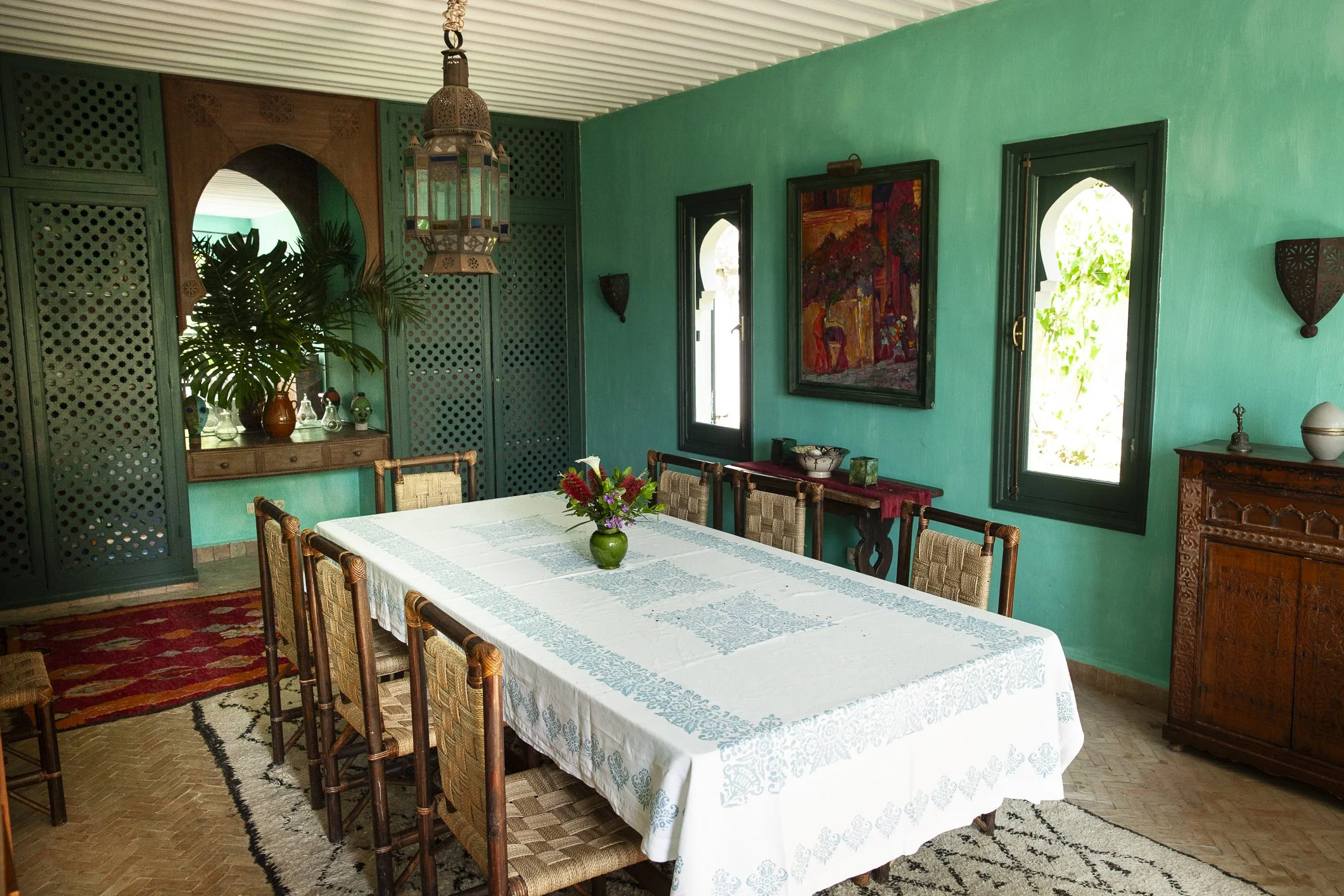 A dining room with a rectangular table covered by a white tablecloth, surrounded by wooden chairs with woven seats. There is a vase with flowers on the table. The room has turquoise walls, two arched windows, and wall-mounted decorative arts. A painting hangs on the wall, and there are various decorative objects and furniture including a wooden cabinet and a side table.