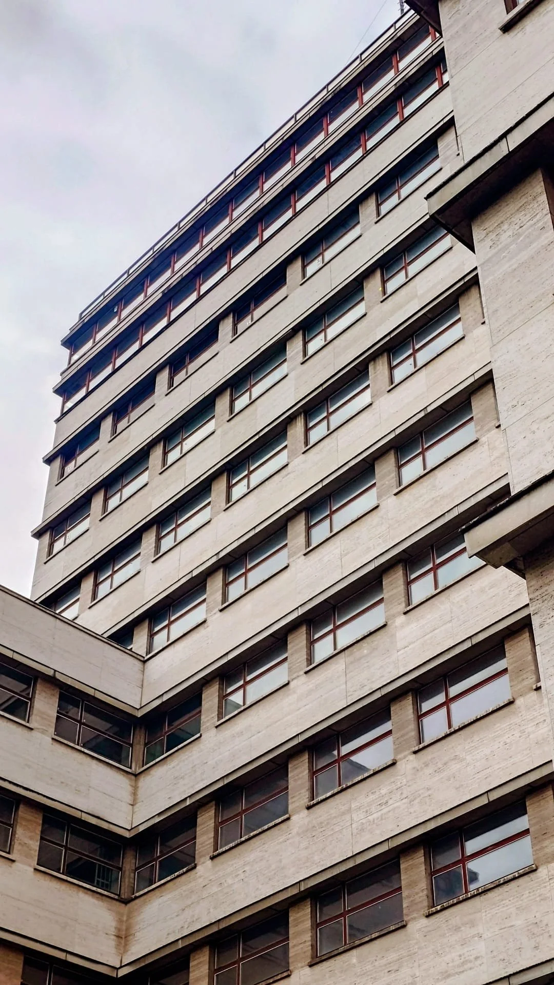 A tall modern building with multiple floors, beige facade, numerous rectangular windows with dark frames, and visible horizontal lines on the exterior wall, viewed from below against a cloudy sky.