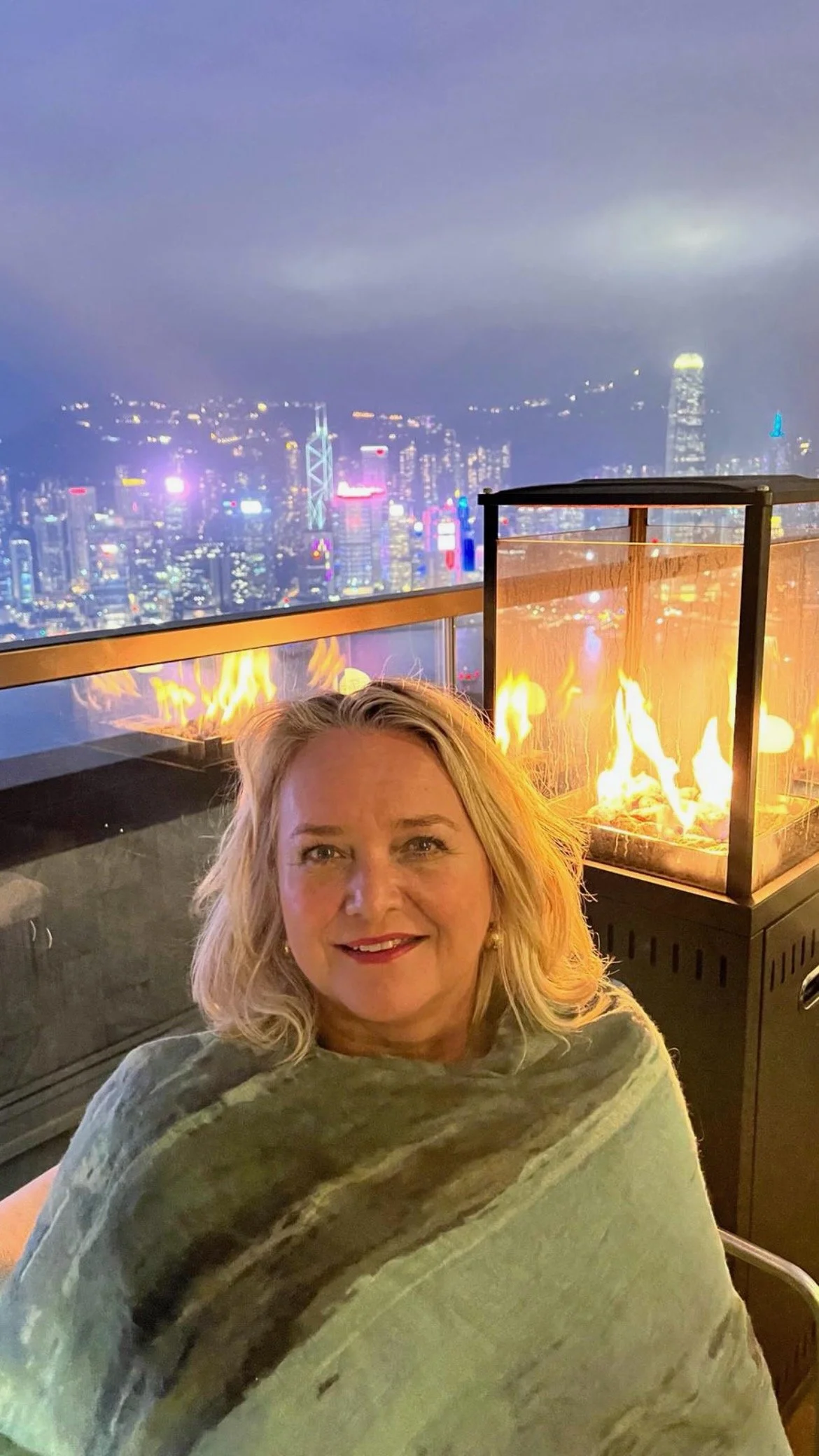 A woman sitting near a firepit with a city skyline at night in the background.