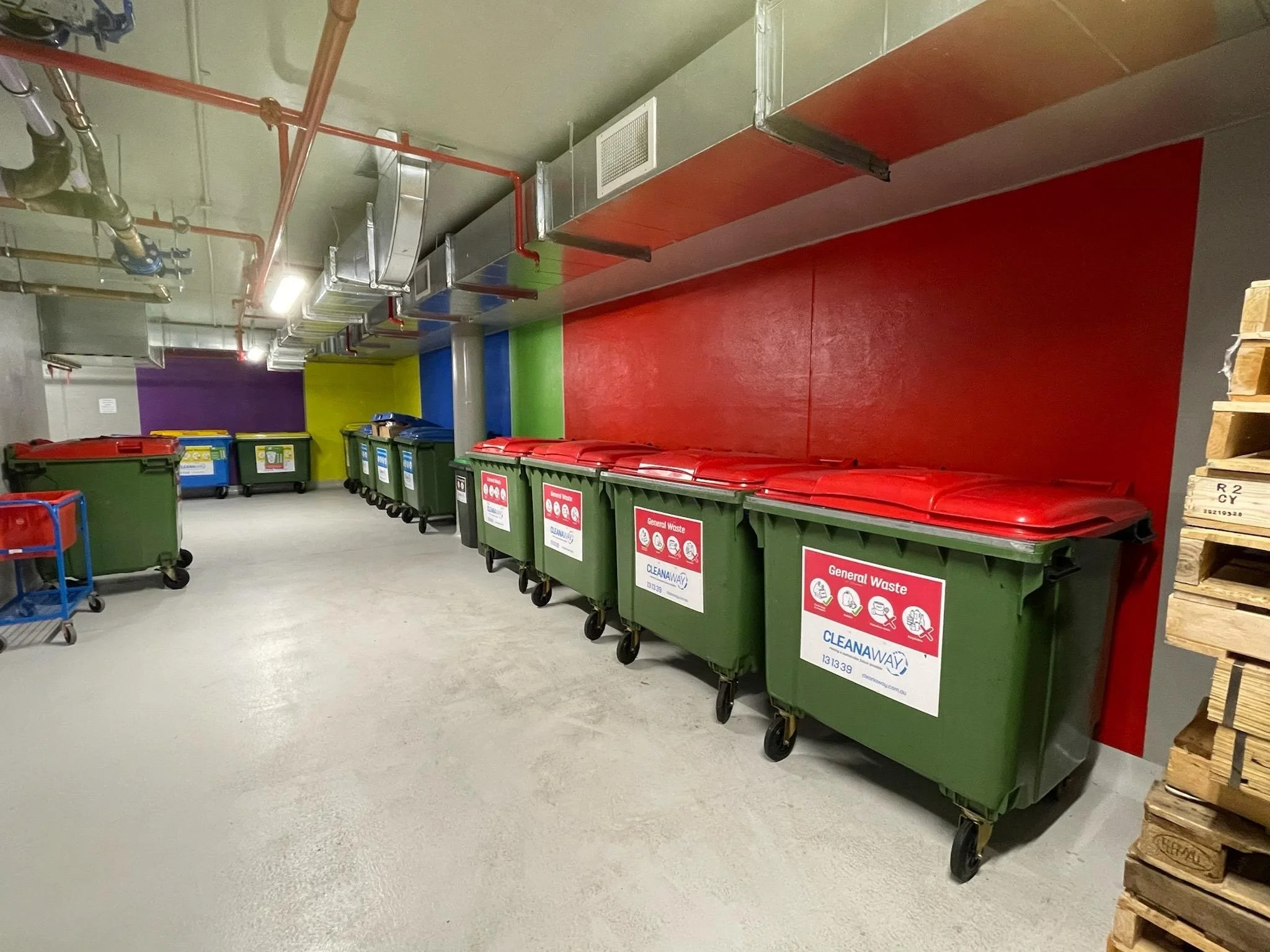 A row of green waste bins with red lids labeled 'General Waste' lined up against a colorful wall with painted sections in purple, yellow, blue, green, and red in an indoor trash disposal area.