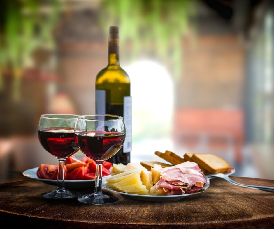 A table with two glasses of red wine, a plate of sliced tomatoes, cheeses, and meats, with a bottle of wine in the background.