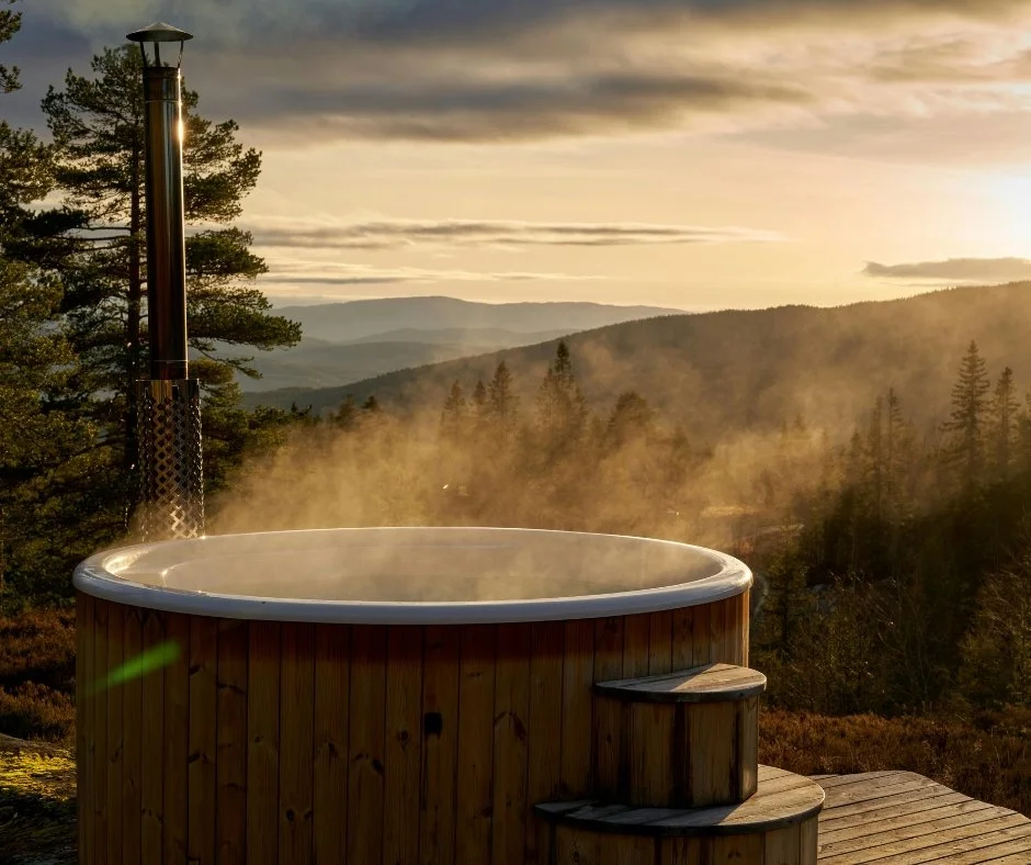 Wooden hot tub on a deck overlooking a forested landscape at sunset with mountains in the background.