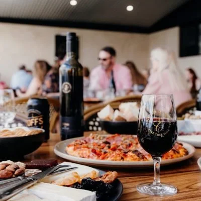 A glass of red wine, a pizza on a plate, and a charcuterie board with cheese and meats on a dining table in a restaurant.