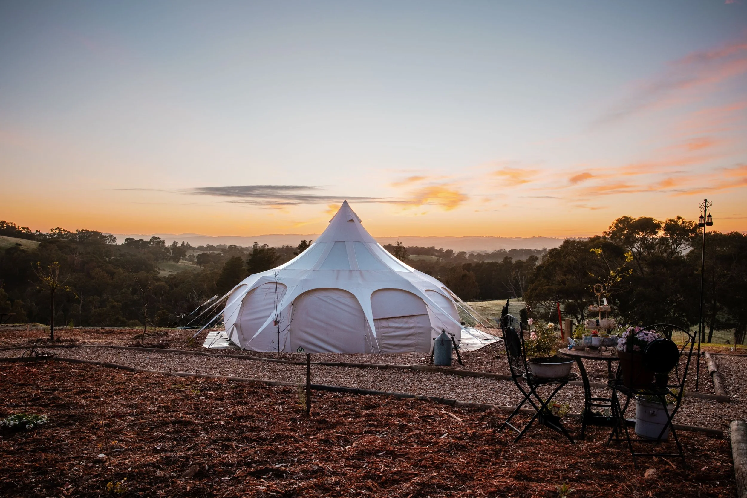 A large white tent with a peaked roof in a desert landscape at dusk, with steps leading to the entrance and trees in the background.