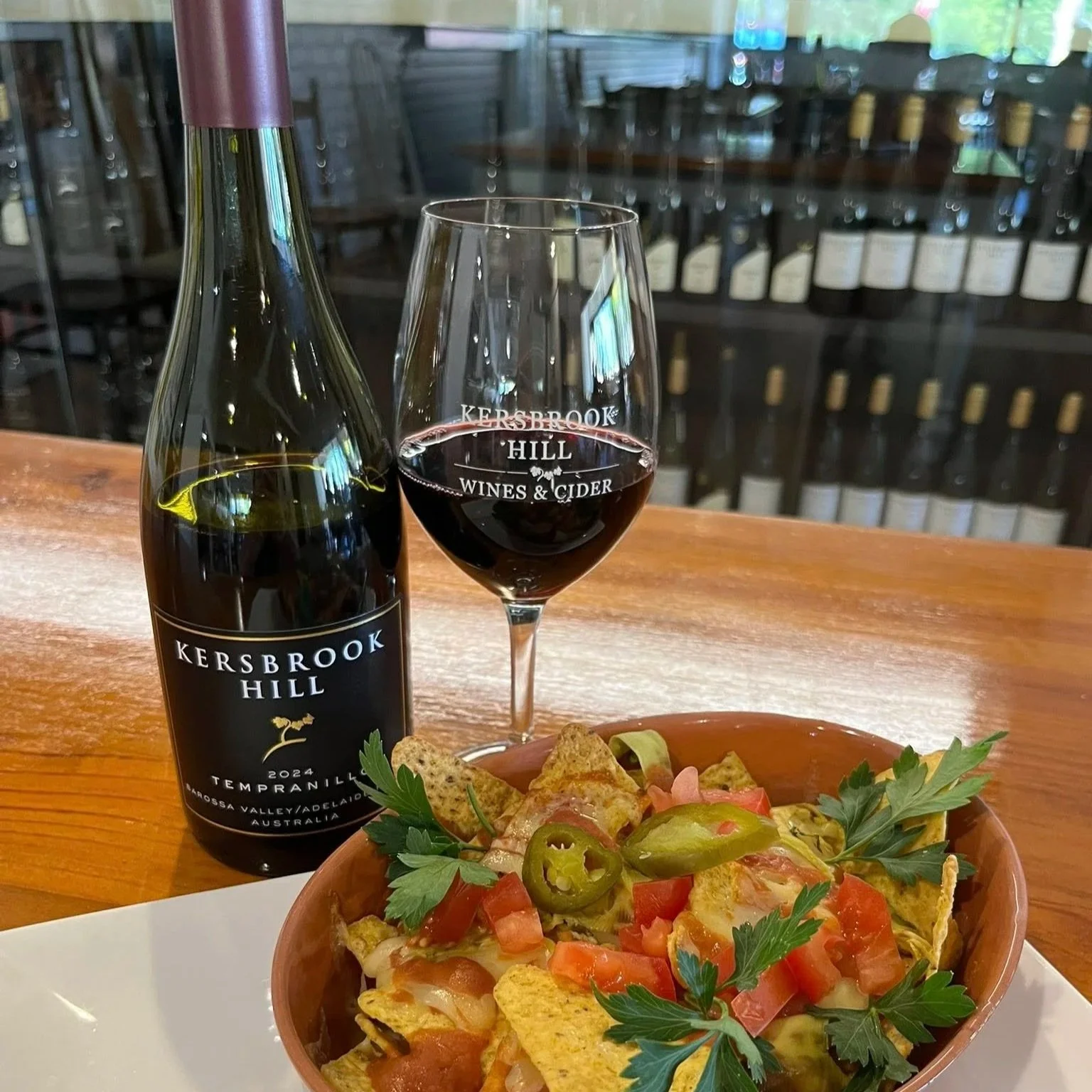 A bottle of Kersbrook Hill Tempranillo red wine, a glass of red wine, and a bowl of nachos topped with tomatoes, jalapenos, cheese, and herbs on a wooden table.