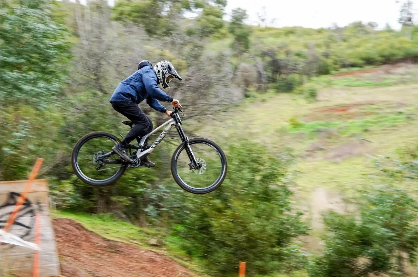 A mountain biker in a blue jacket and black pants riding a downhill trail on a gray mountain bike with full suspension, in a forested area with green foliage.