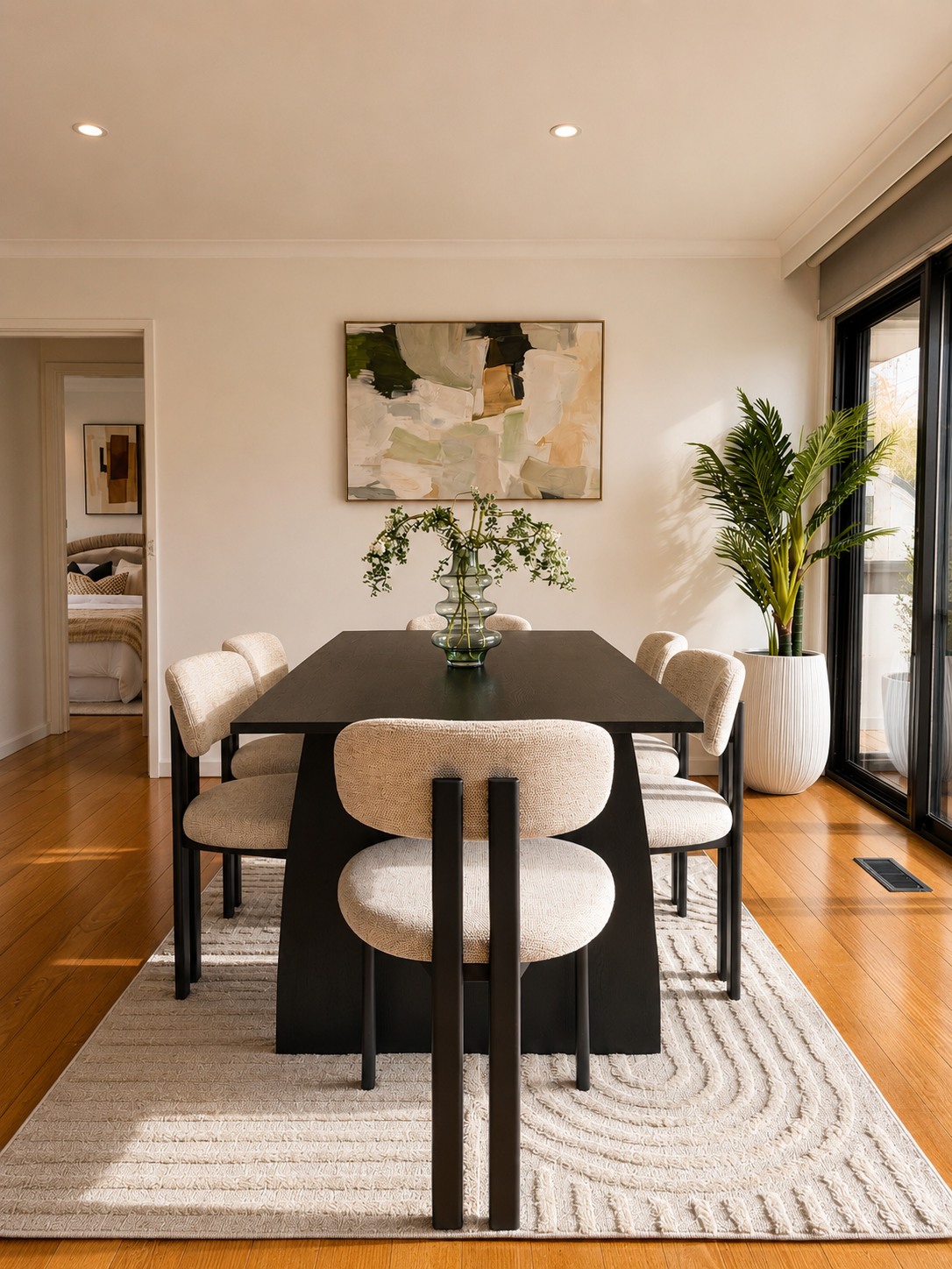 A living room with a large black vase holding pink flowering branches on a wooden sideboard, with a sculpture of a horse and books nearby. There is a chair and glass doors leading outside to a backyard with a pool and greenery.