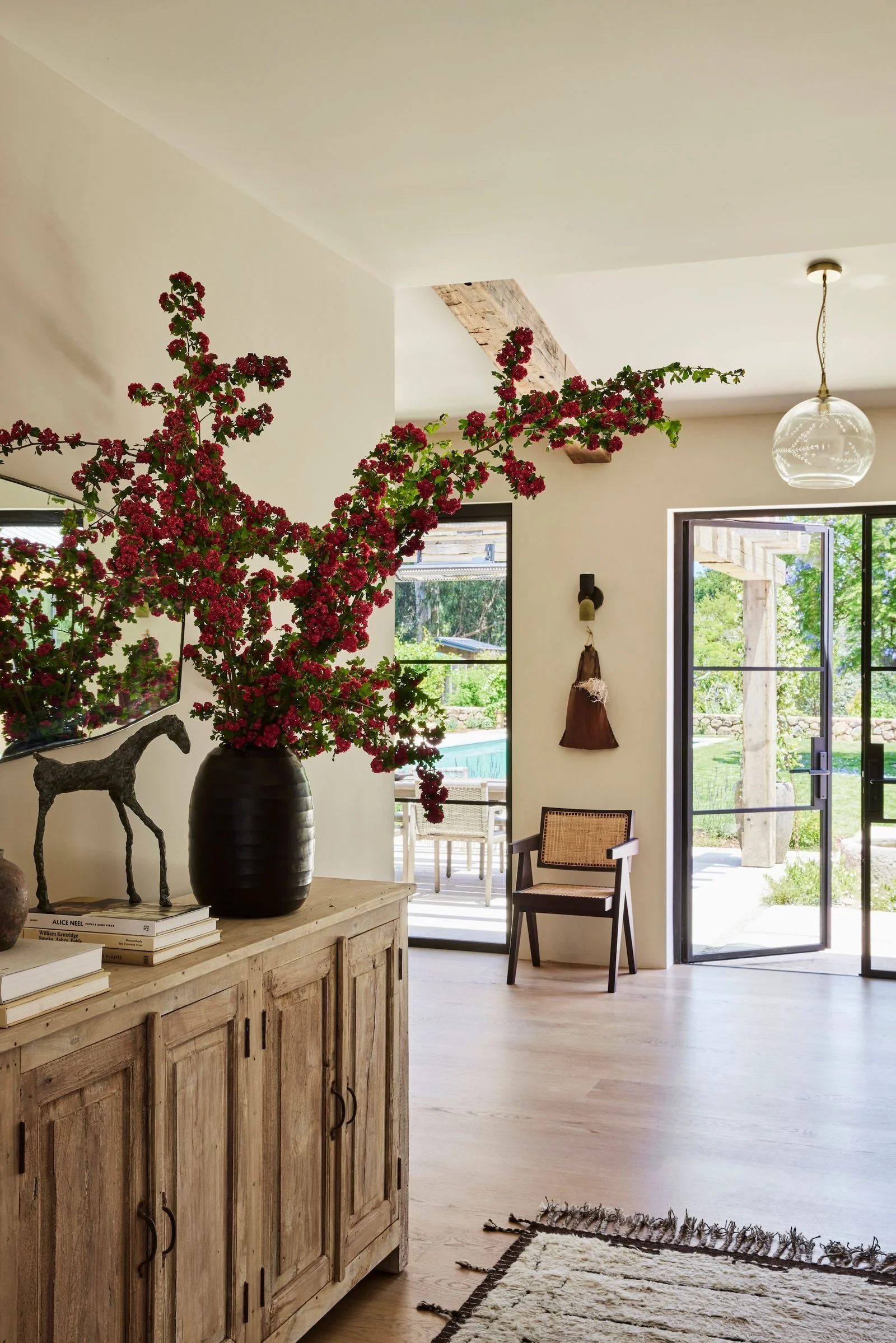 A living room with a large black vase holding pink flowering branches on a wooden sideboard, with a sculpture of a horse and books nearby. There is a chair and glass doors leading outside to a backyard with a pool and greenery.