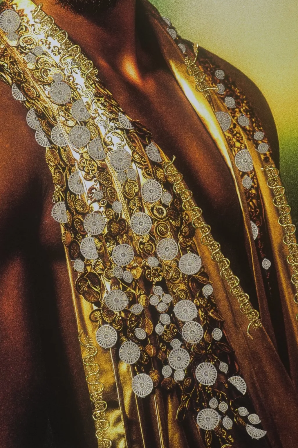 Close-up of a person wearing ornate gold and silver jewelry, including multiple necklaces with intricate patterns and circular medallions, and a richly embroidered garment with gold and black details.