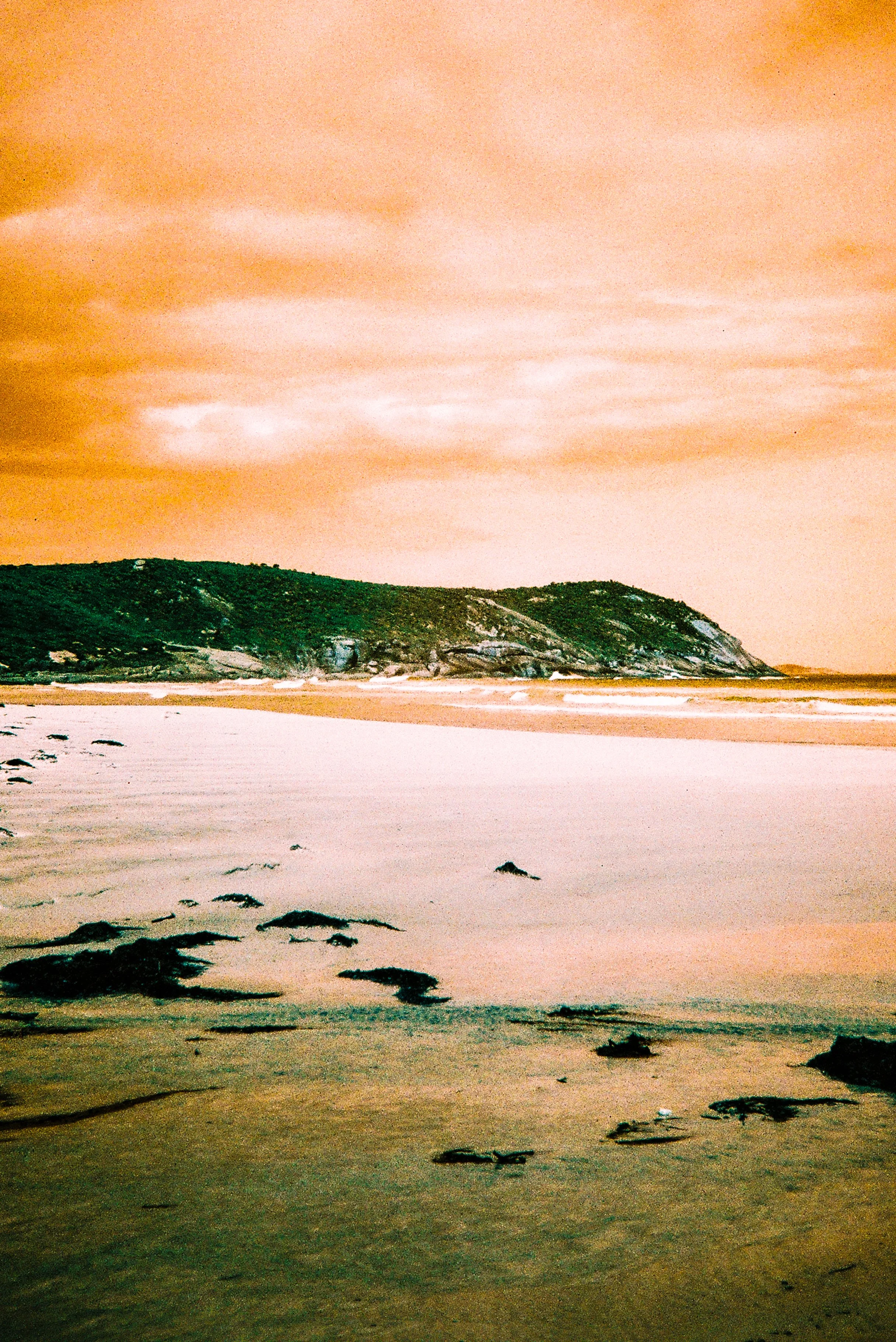 Beach scene with rocky shoreline, a green hill in the distance, and a sky with orange clouds.