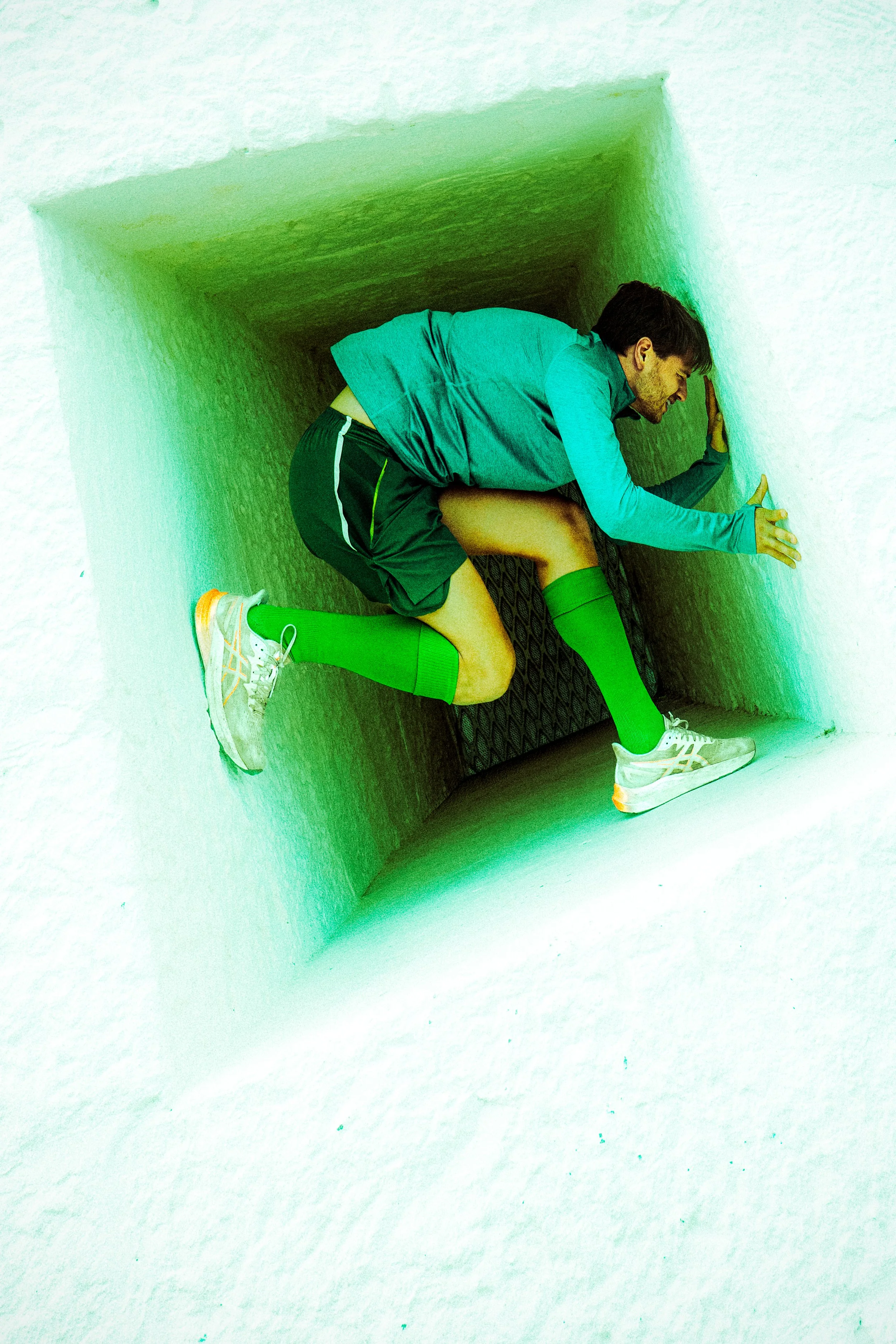 A man in athletic clothing crouches in a small, illuminated, square room with white walls, pushing against one wall with his hands.
