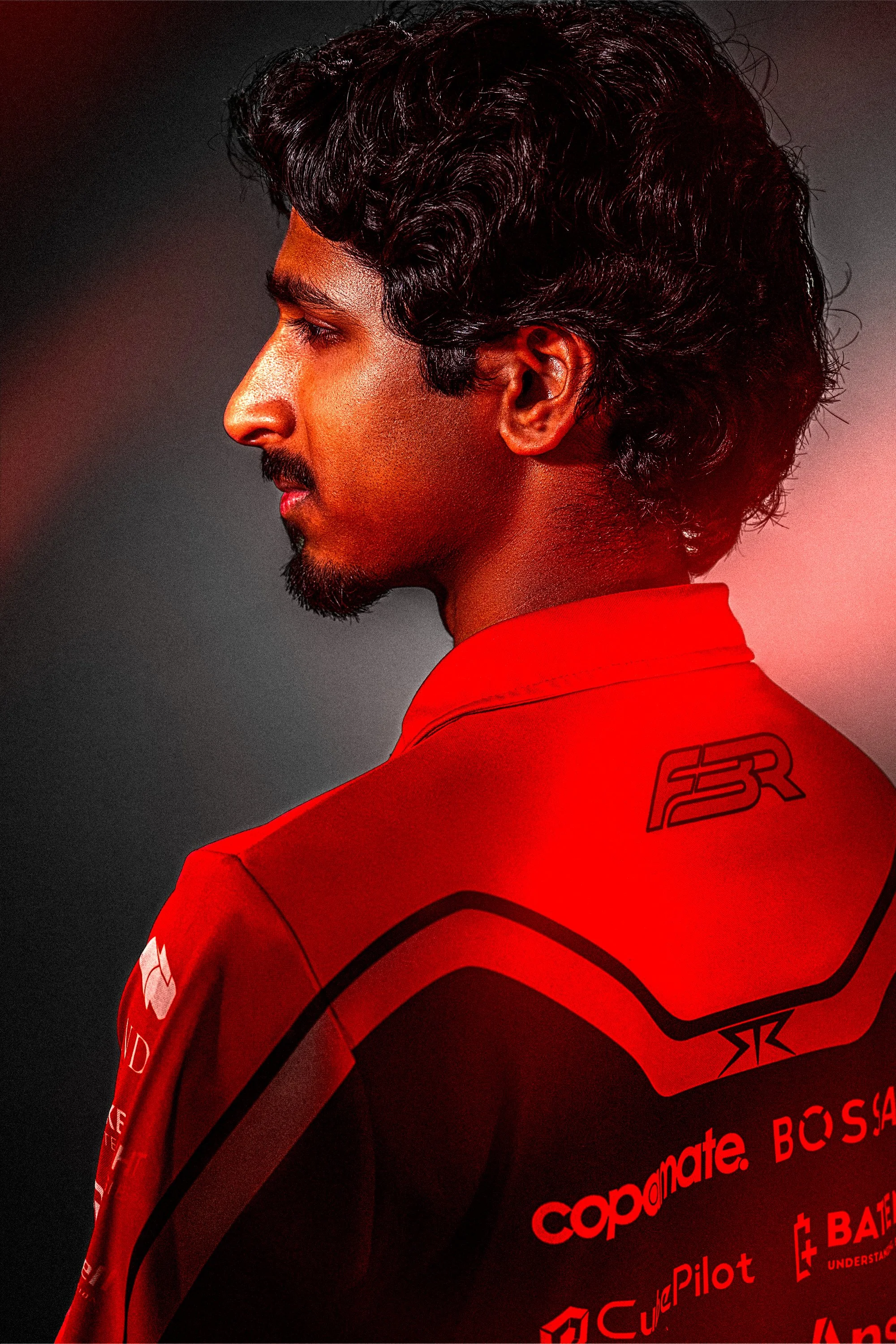 A young man with wavy black hair and a beard is shown from the side, wearing a red sports jersey with black and white details and text logos on the back. The background is dark.