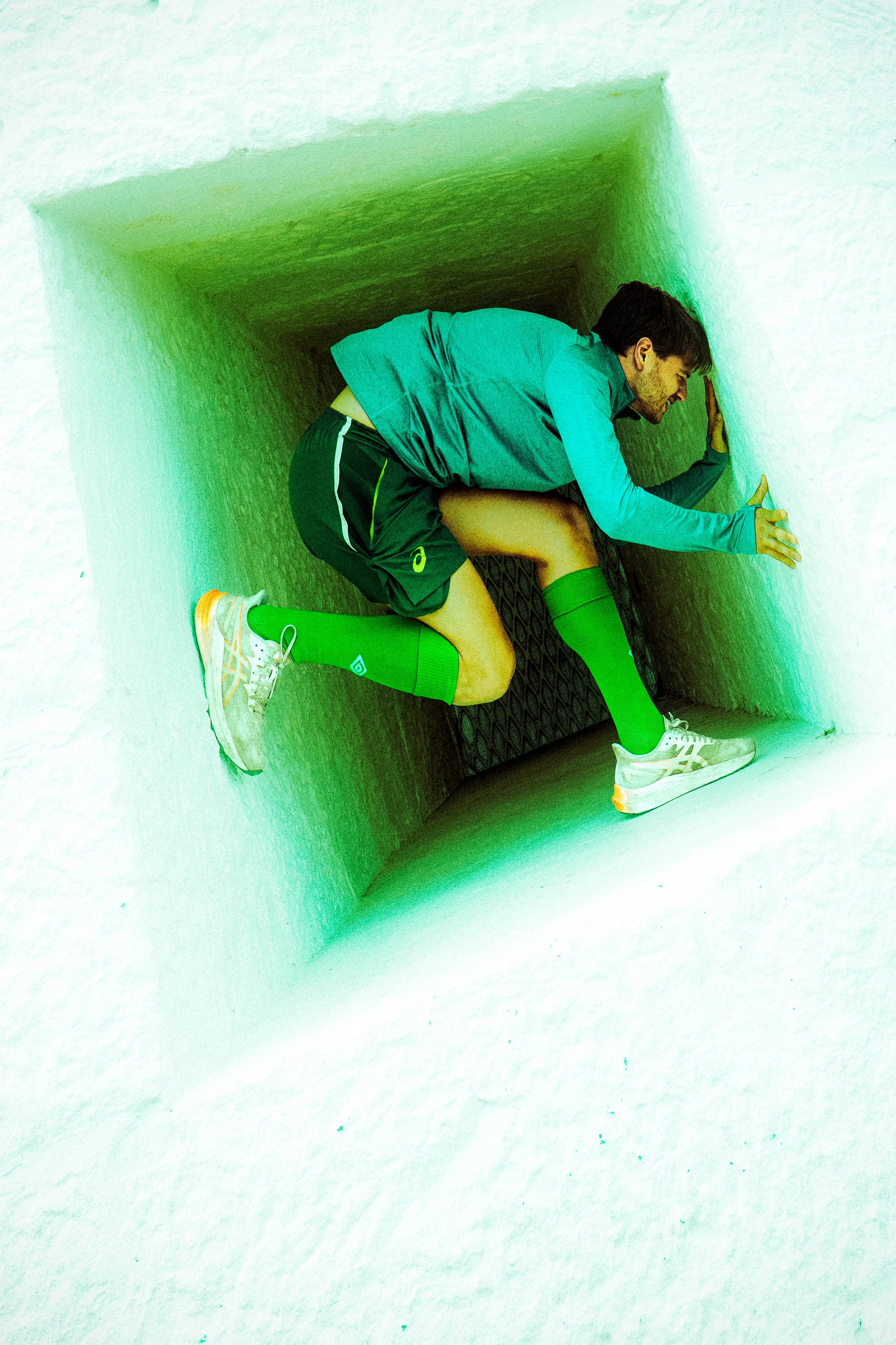 A man crouching inside a square vent or duct, looking downward with hands on the sides of the opening, wearing a turquoise shirt, black shorts, green knee-high socks, and sneakers.