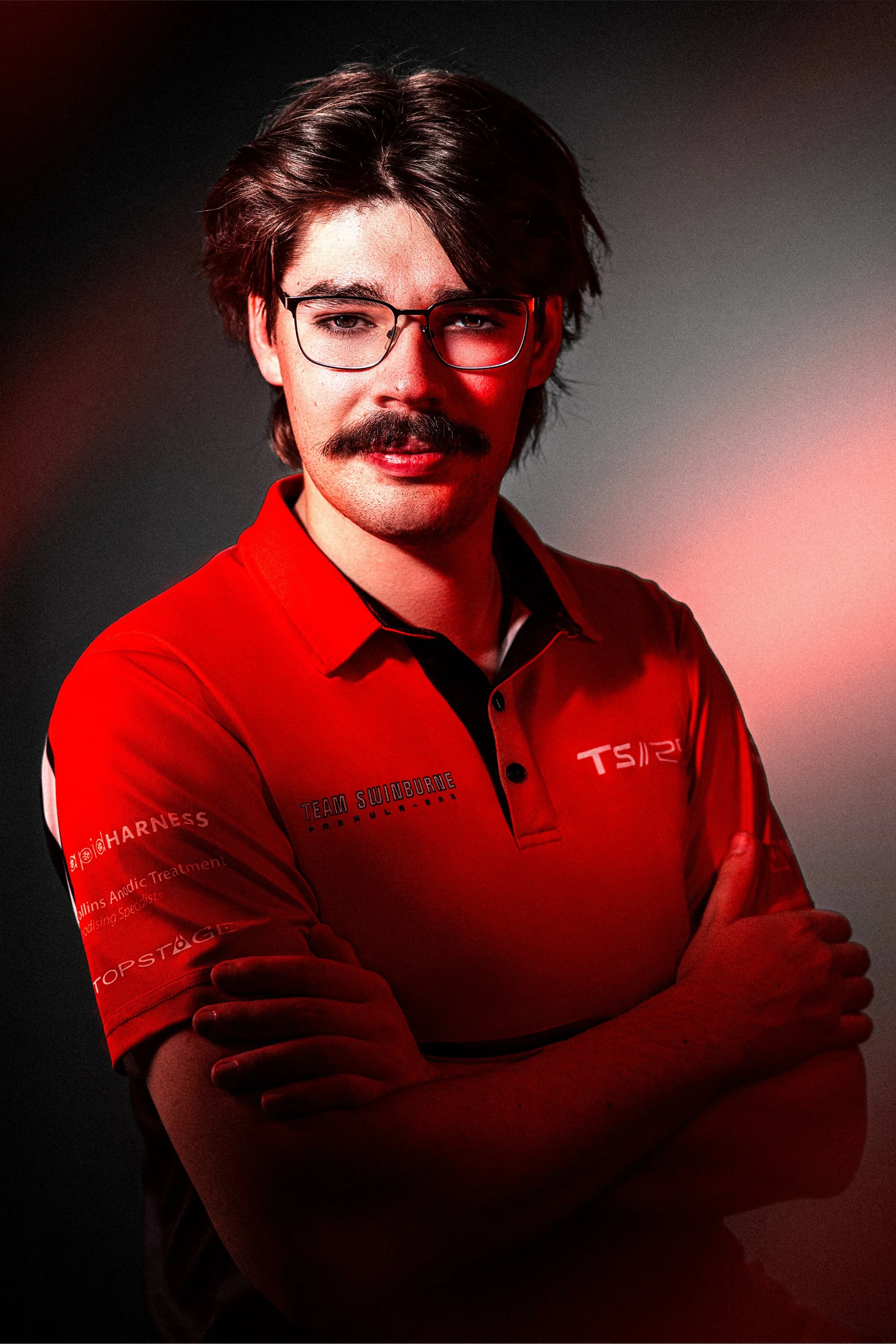 A man with dark brown hair, glasses, and a mustache is wearing a red team uniform with patches and logos. He has his arms crossed and is looking at the camera in a professional studio setting with a dark background and red lighting.