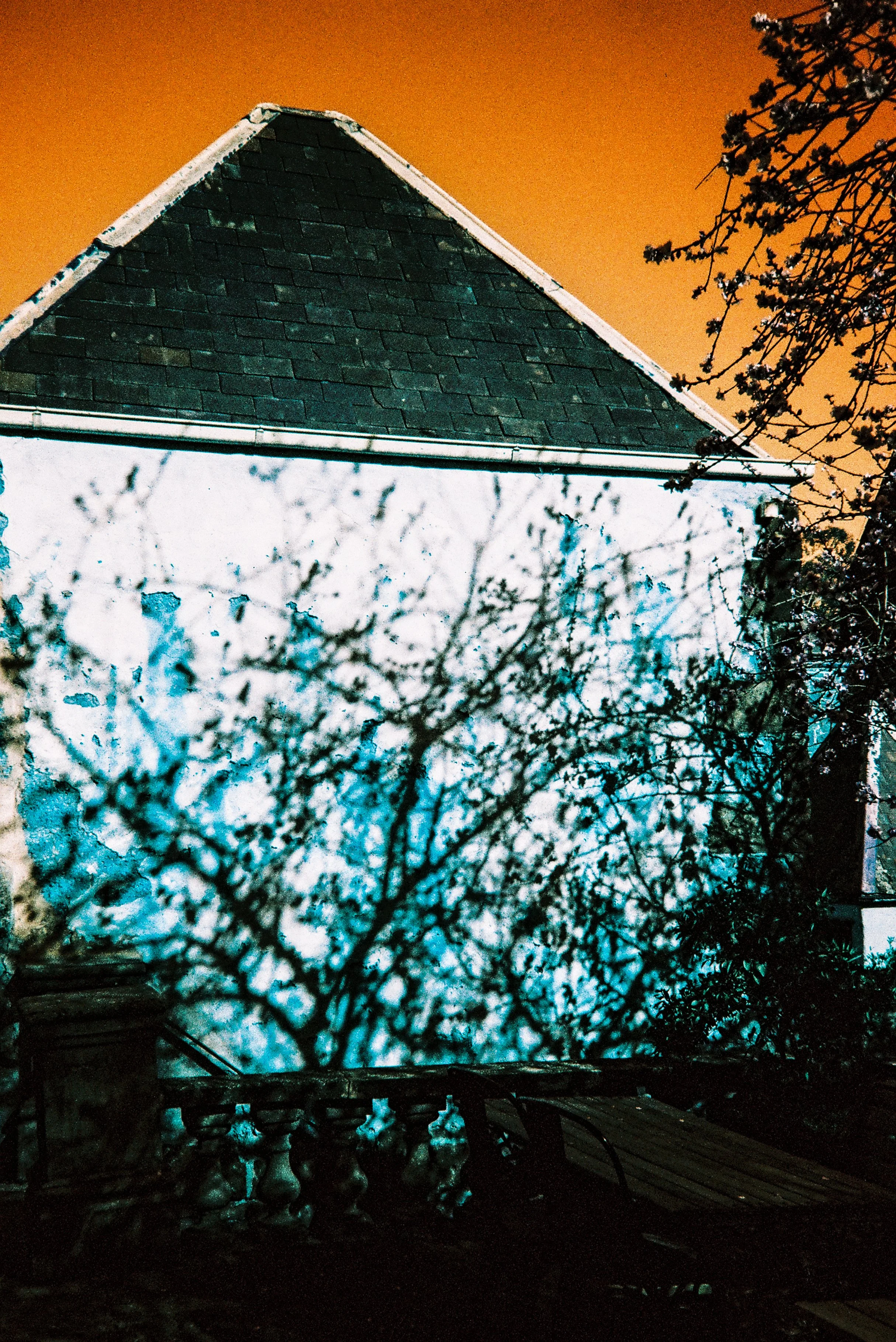 Nighttime view of a house with a triangular roof, partially obscured by leafless tree branches, against an orange sky.