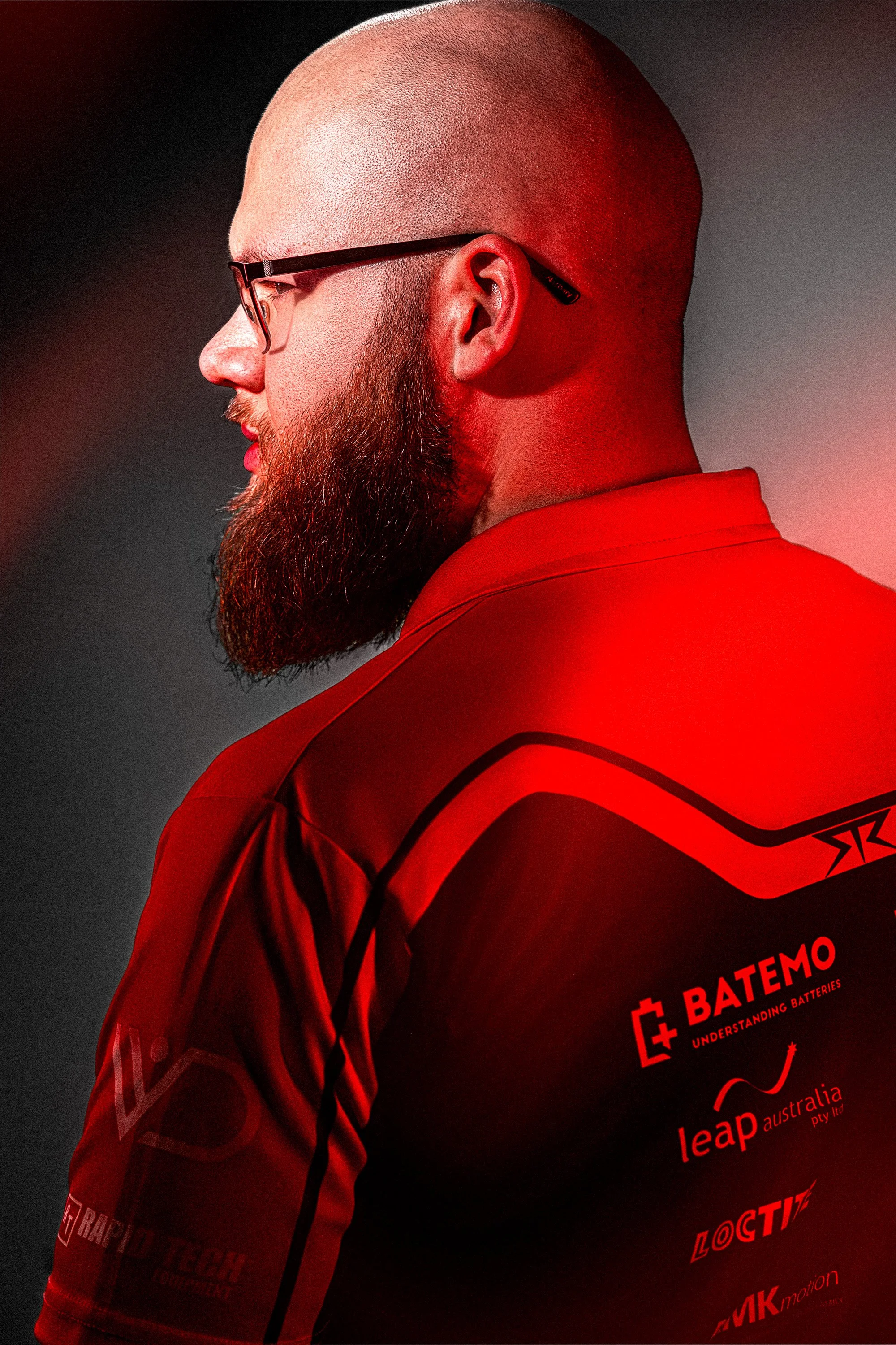Side profile of a man with a shaved head and beard wearing glasses and a red sports jersey with logos.