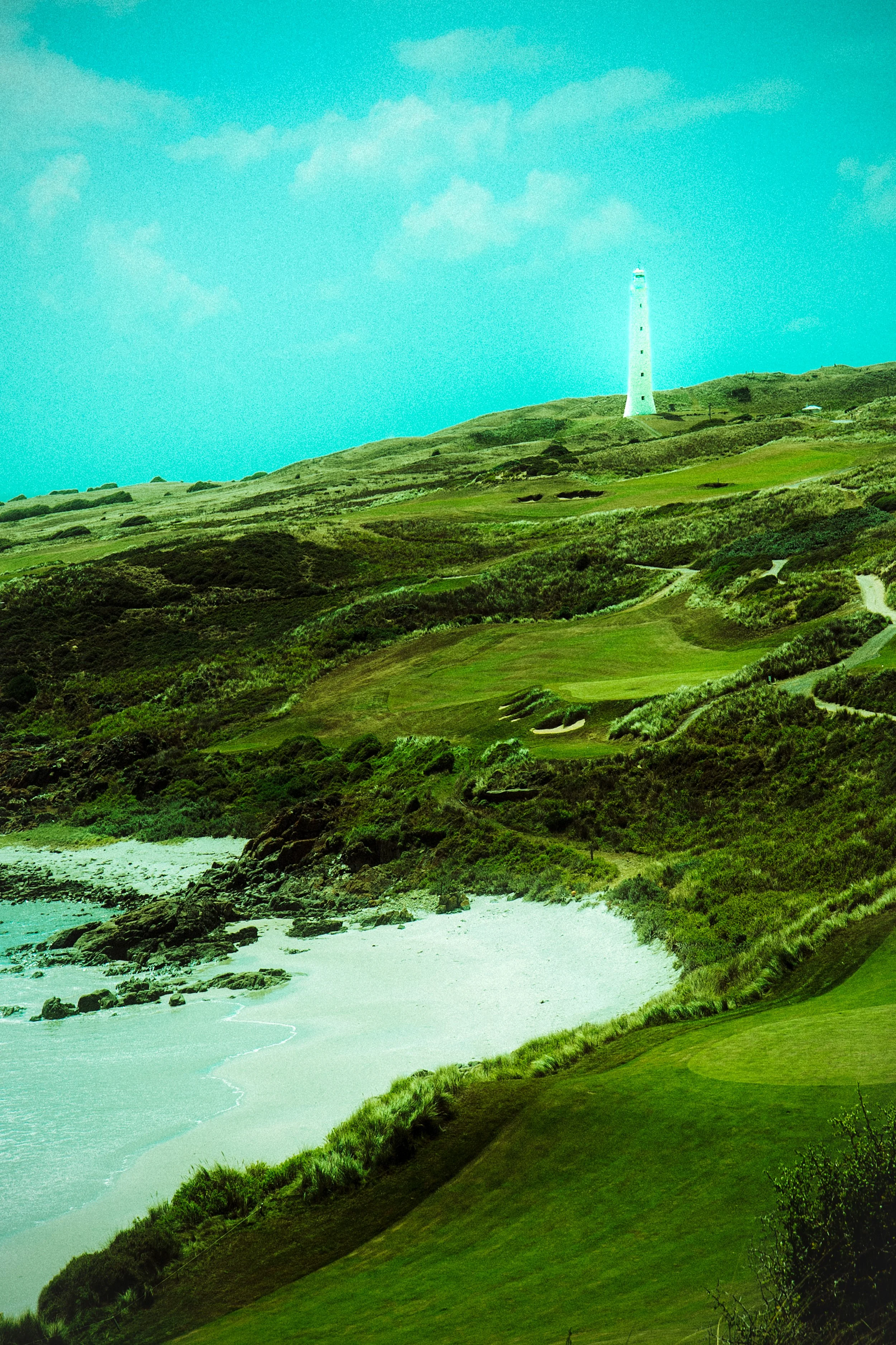 Scenic view of a coastal golf course with lush green fairways, a sandy beach, and a lighthouse on the hilltop under a blue sky with scattered clouds.