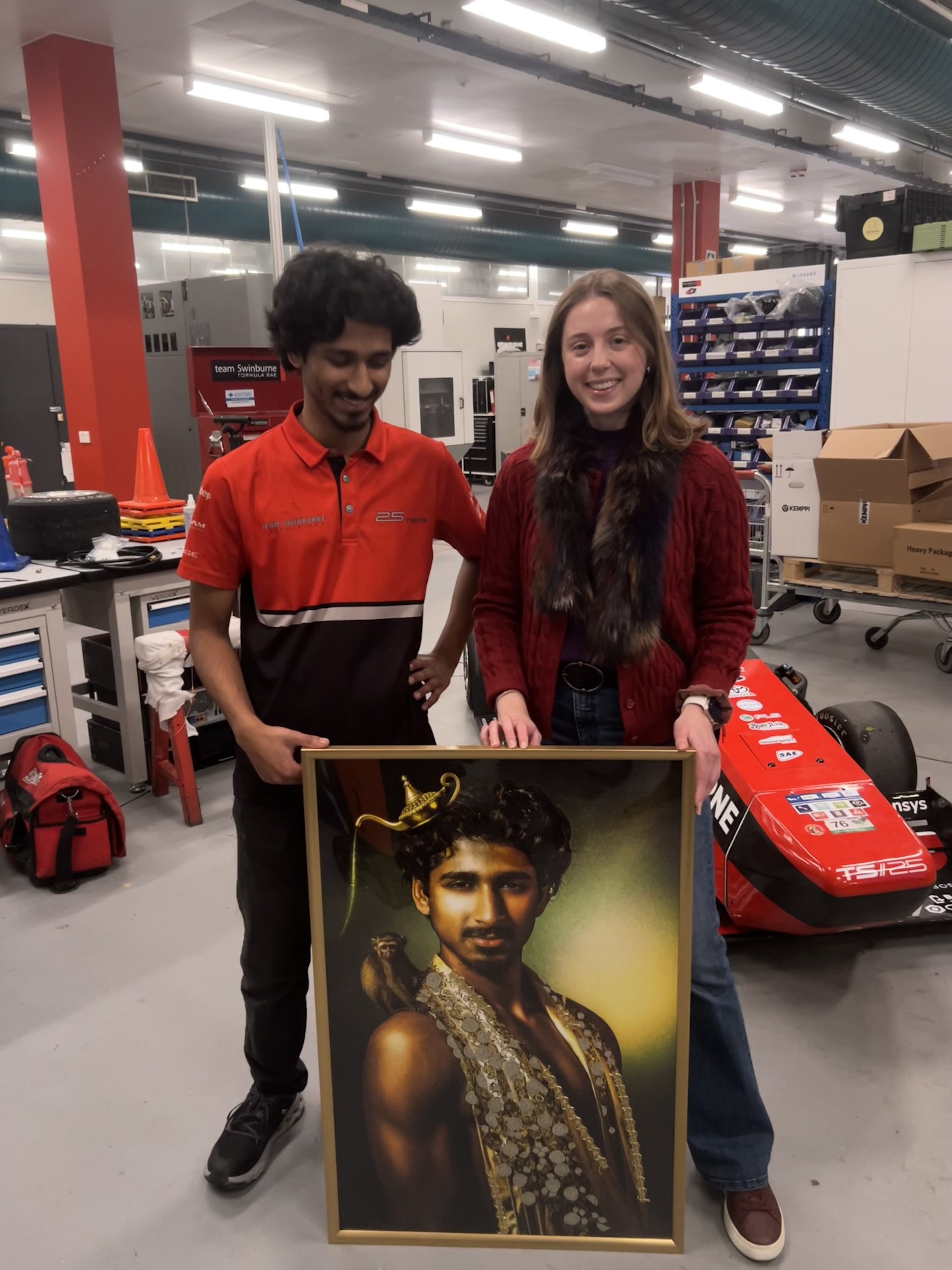 Two people standing inside a workshop or garage, holding a large framed portrait of a woman wearing jewelry and traditional attire. Shelves and boxes are visible in the background.