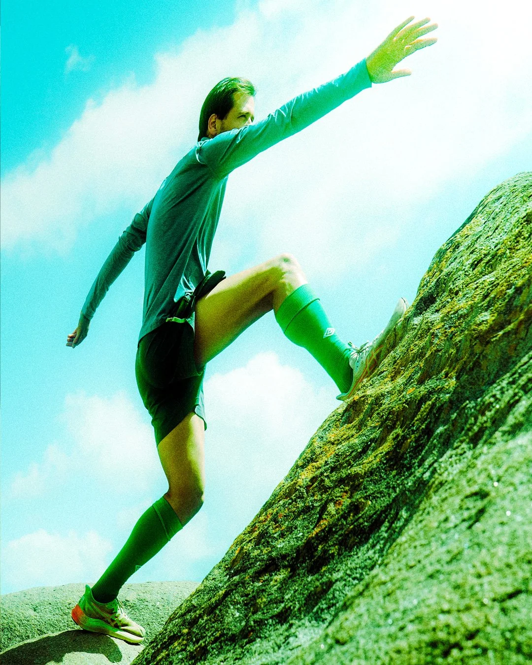 Person climbing a green and gray rocky slope outdoors under a blue sky.