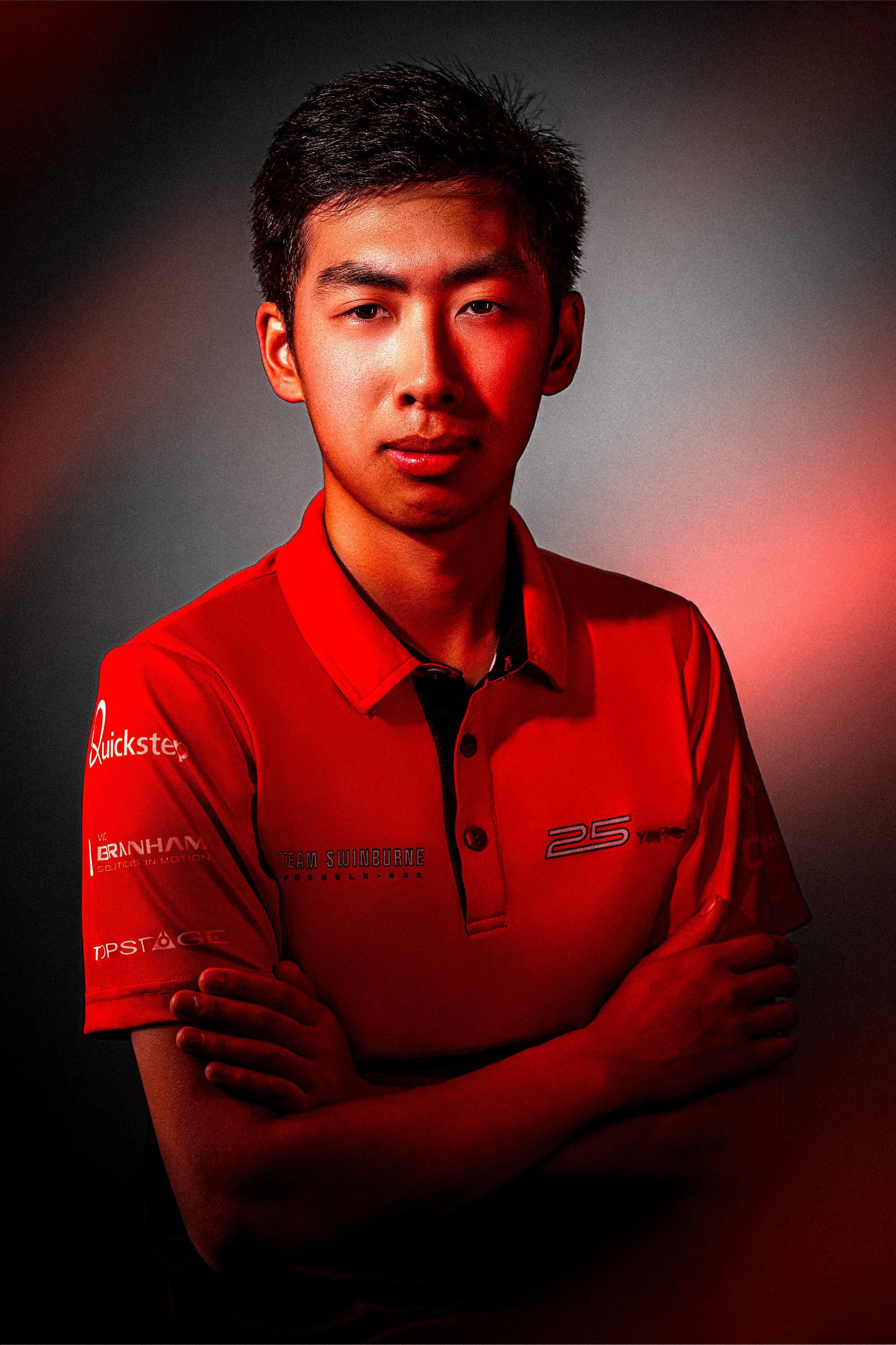 Young male racing driver in red team uniform with arms crossed, standing against a dark background with red lighting accents.