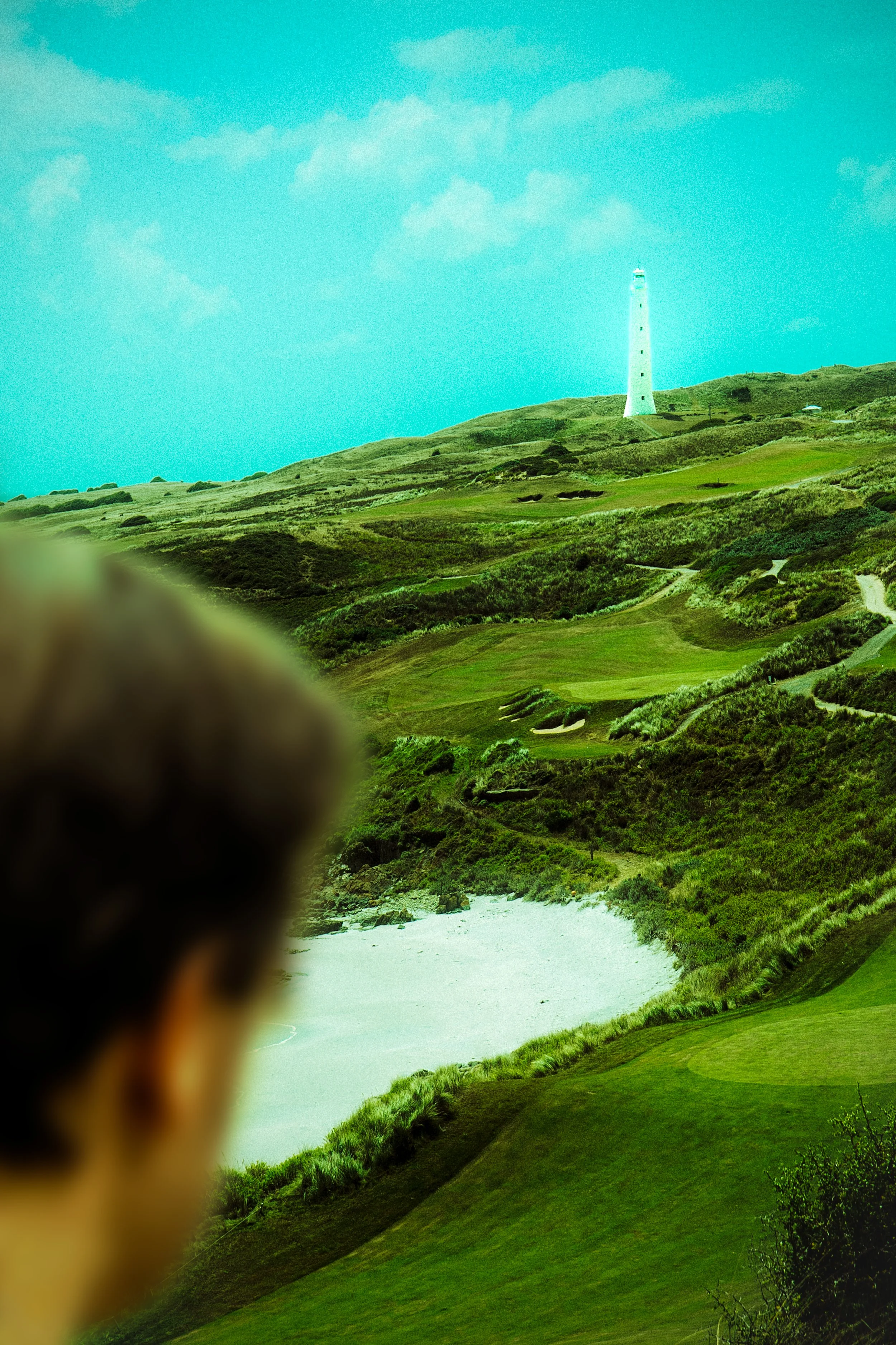A scenic view of a lighthouse on a grassy hillside near the ocean, with rolling green hills and a sandy beach in the foreground.