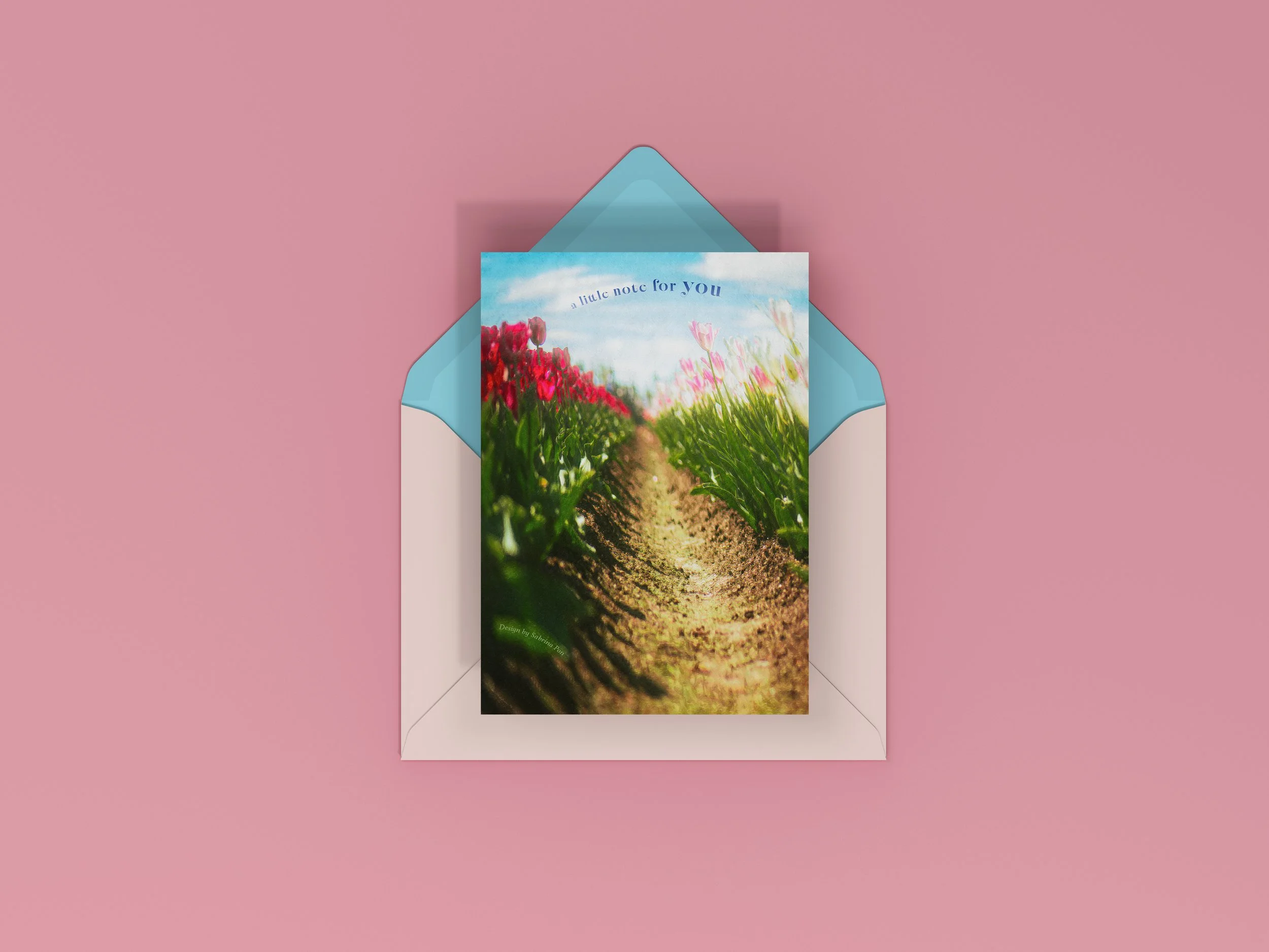 A greeting card with a photograph of a flower field and a dirt path, placed inside a light blue envelope on a pink background.