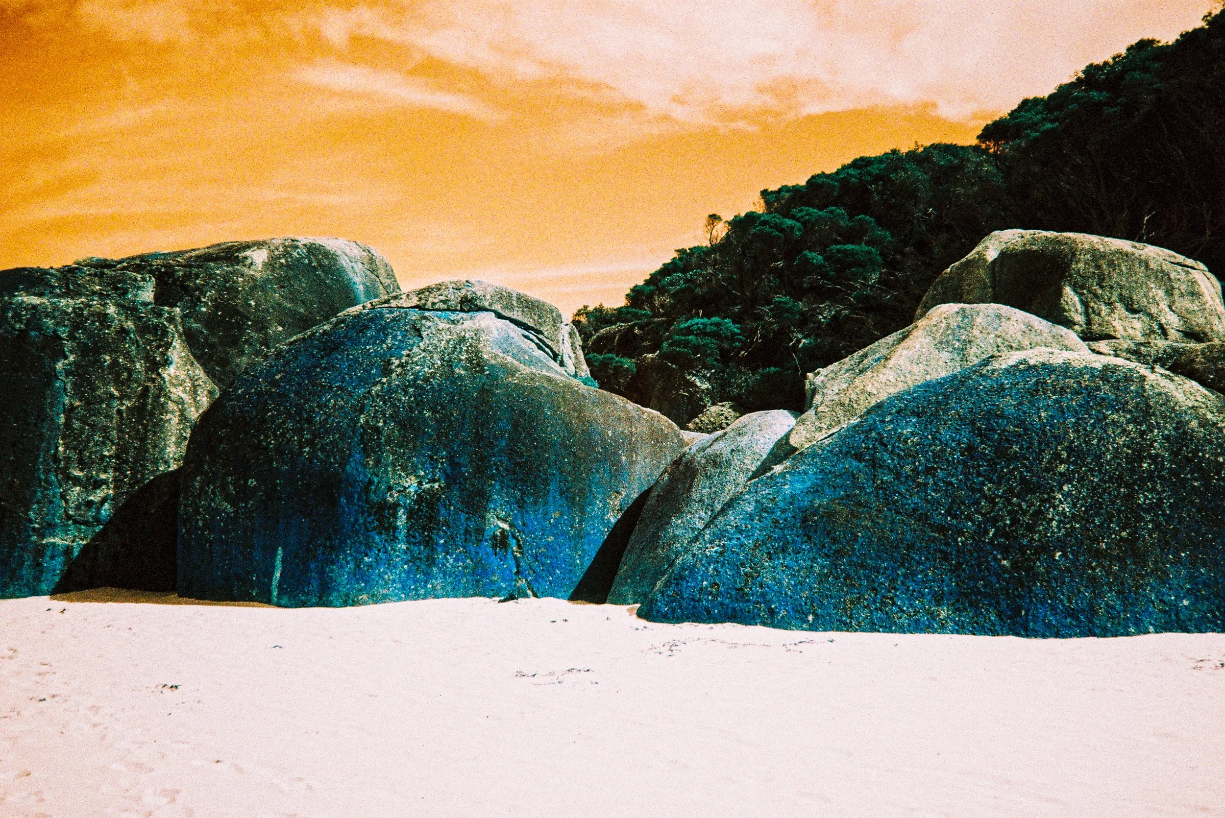 Large boulders on sandy beach with trees and a sunset sky in the background.