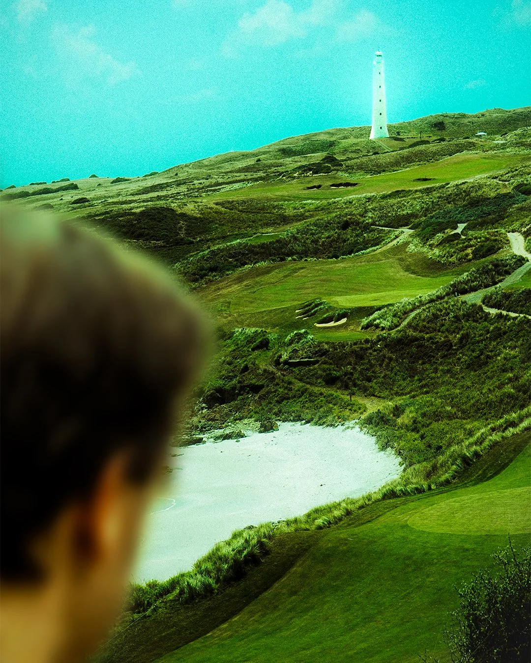 A person with dark hair watching a landscape with green rolling hills, a lighthouse on top, and a white sandy beach near the water in the distance.