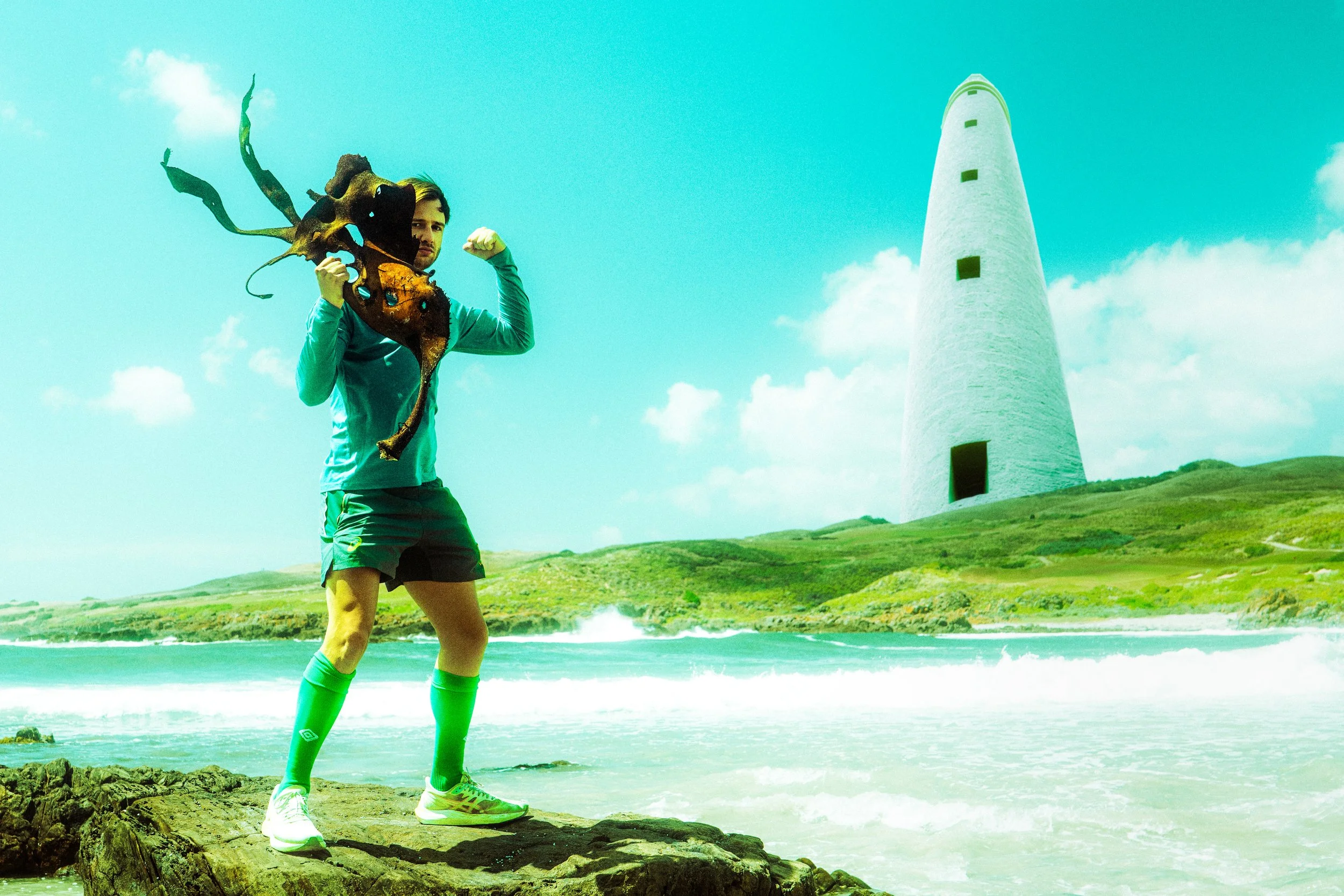 A man in athletic wear standing on a rocky surface at the beach, holding a large, colorful dragon mask and flexing his arm muscles, with a lighthouse and green hills in the background under a blue sky with clouds.