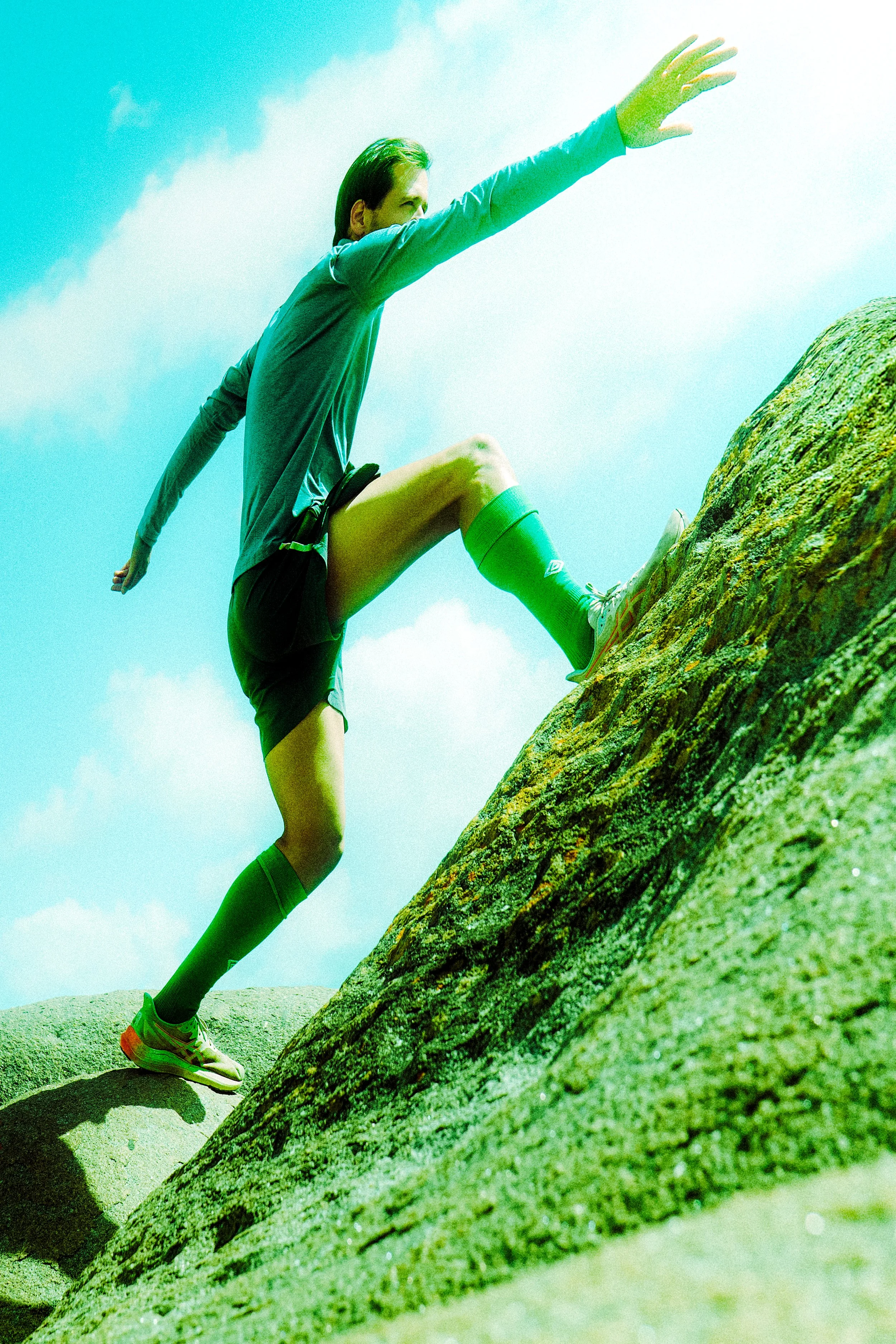 Person climbing a steep, moss-covered rock outdoors on a sunny day.