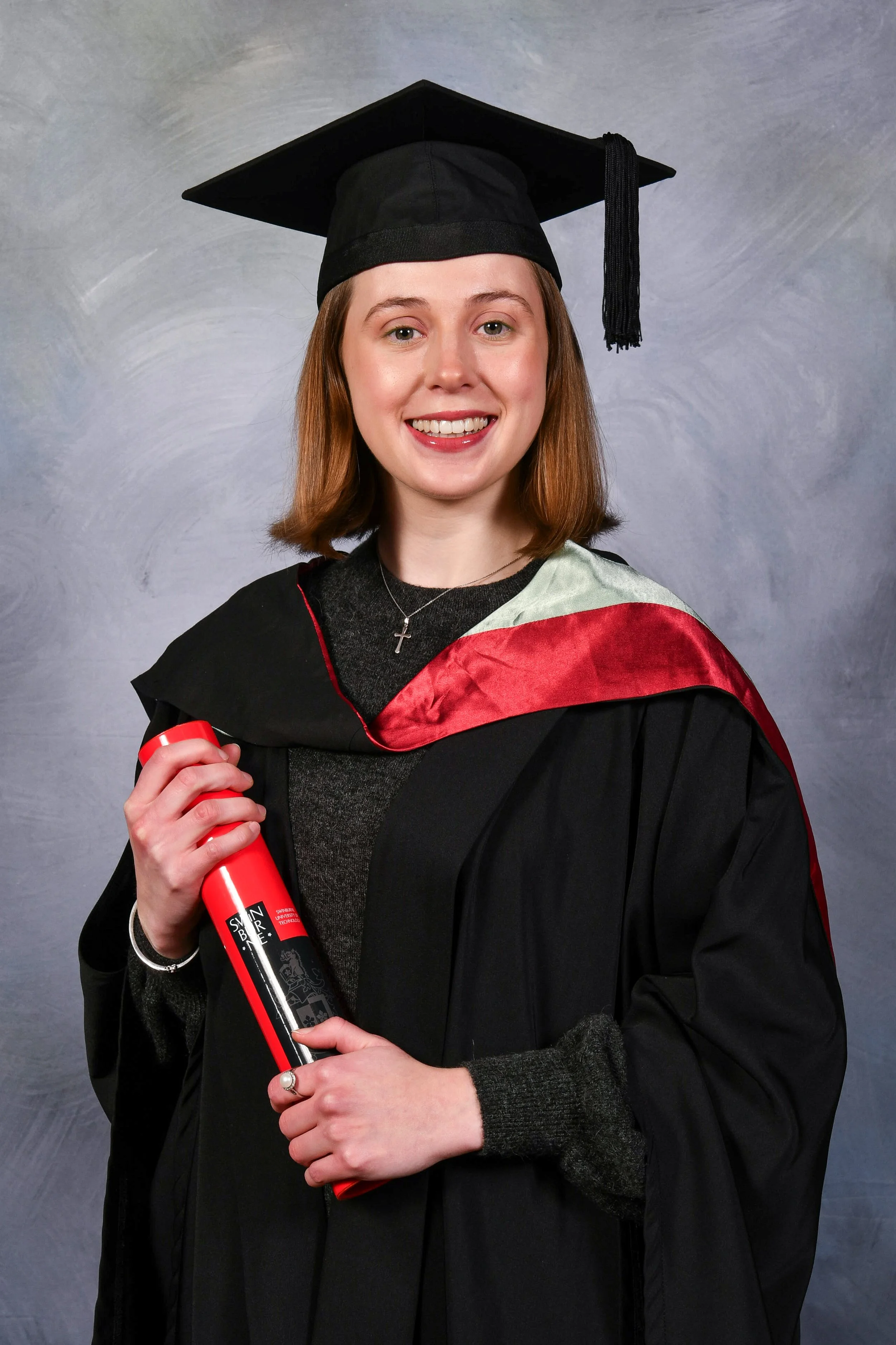 A young woman wearing a black graduation cap and gown, holding a red diploma and smiling at the camera. She is wearing a cross necklace and a multicolored stole.