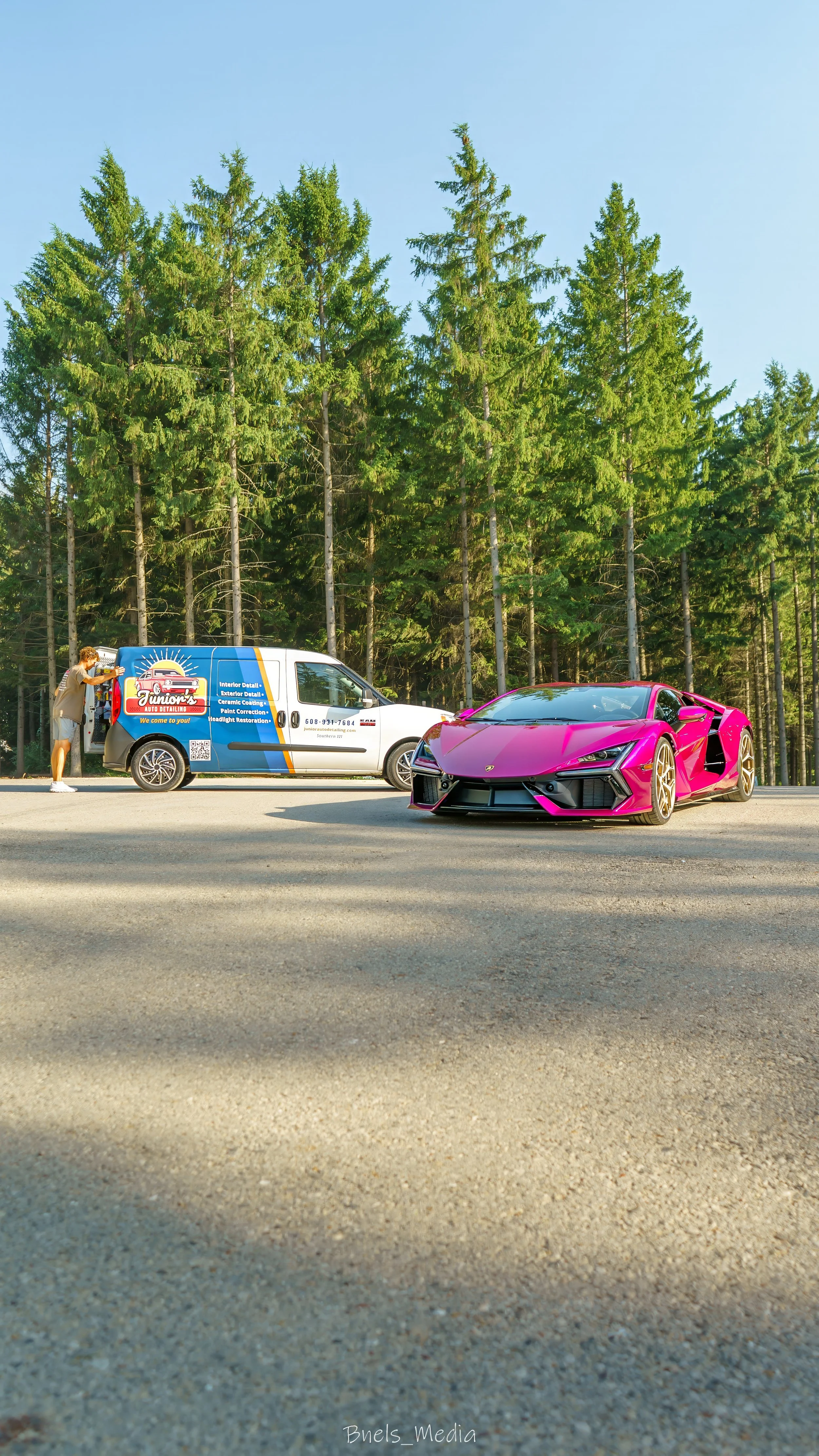 A vibrant pink Lamborghini sports car parked on the side of a road next to a white vehicle with a blue and orange advertisement for auto detailing. A person is standing near the white vehicle, possibly working on or inspecting it. The background features a dense forest of tall green trees under a clear blue sky.