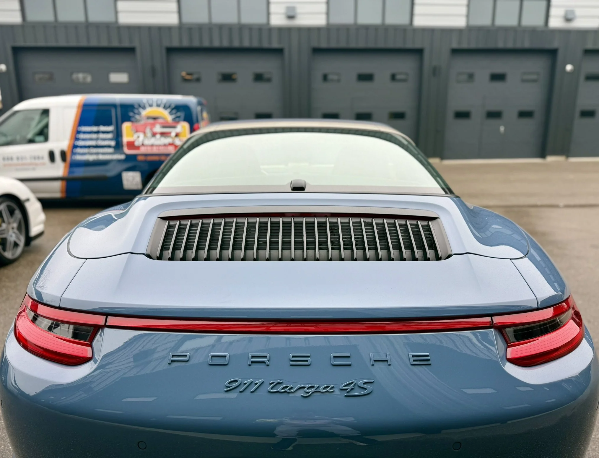 Rear view of a blue Porsche 911 Targa 4S parked in a lot, with a blue vehicle with colorful graphics and a white vehicle on the left in the background, and gray garage doors behind.