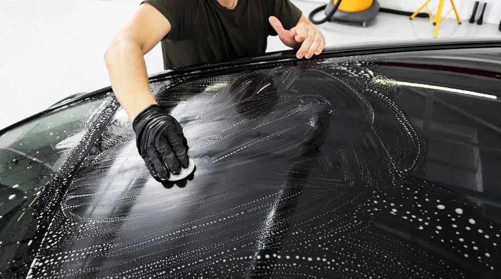 Person cleaning a black car's windshield with soapy water and a scrubber, wearing black gloves.