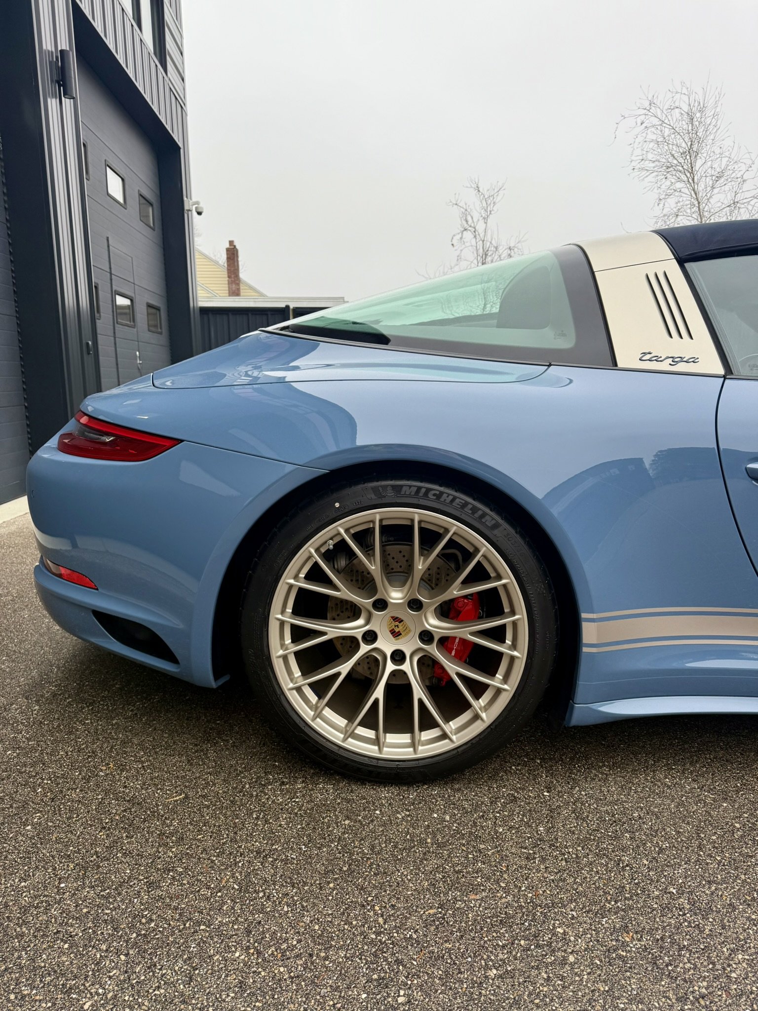 A light blue Porsche Targa convertible car parked outside, showing the rear and part of the side with a focus on the wheel, brake caliper, and rear light.